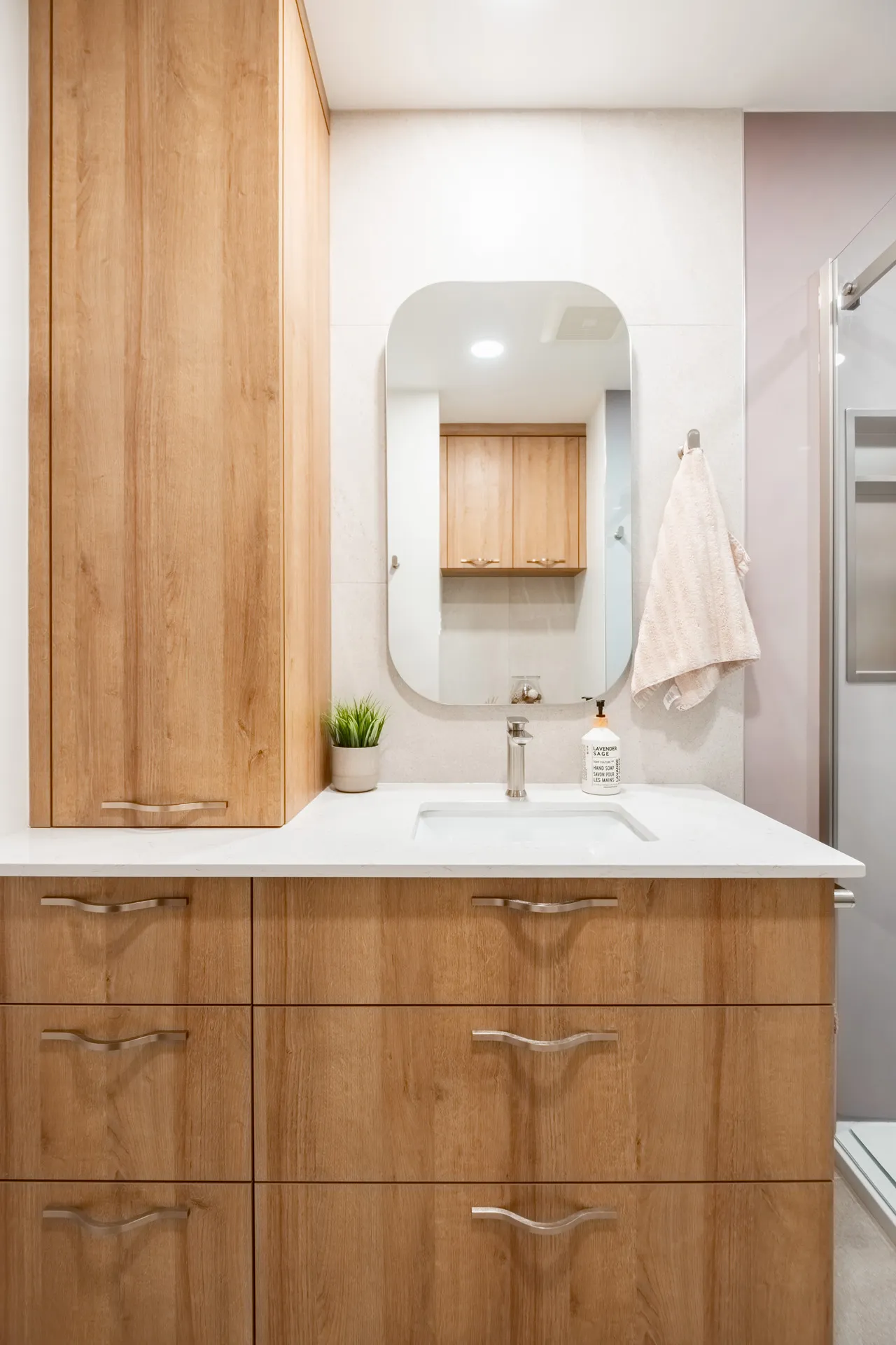 Salle de bain Menuitech avec armoires en bois clair, comptoir blanc, miroir arrondi et accessoires minimalistes