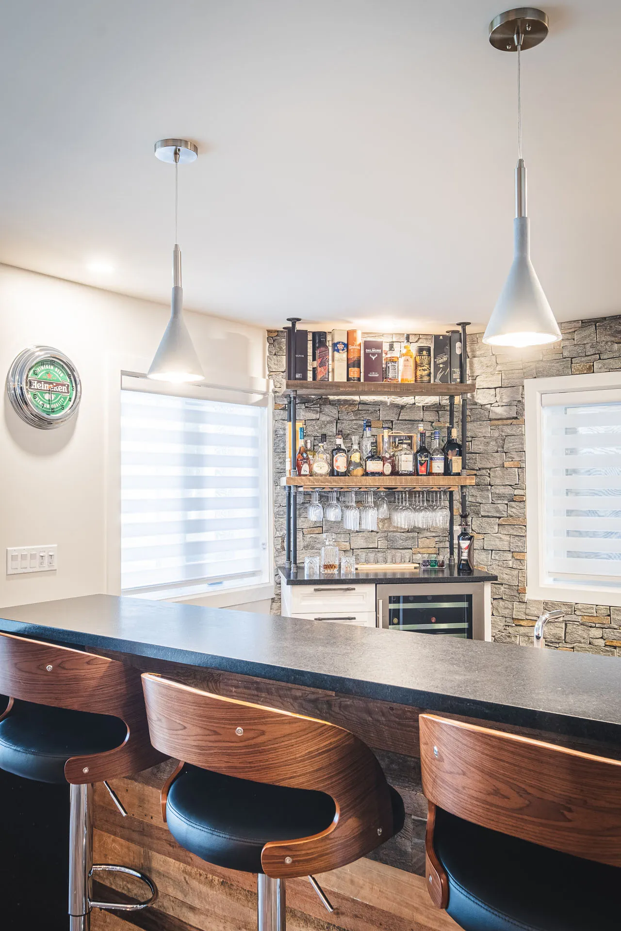 Residential bar with gray island, wood and black leather stools, stone backsplash, metal shelving, white pendant lights