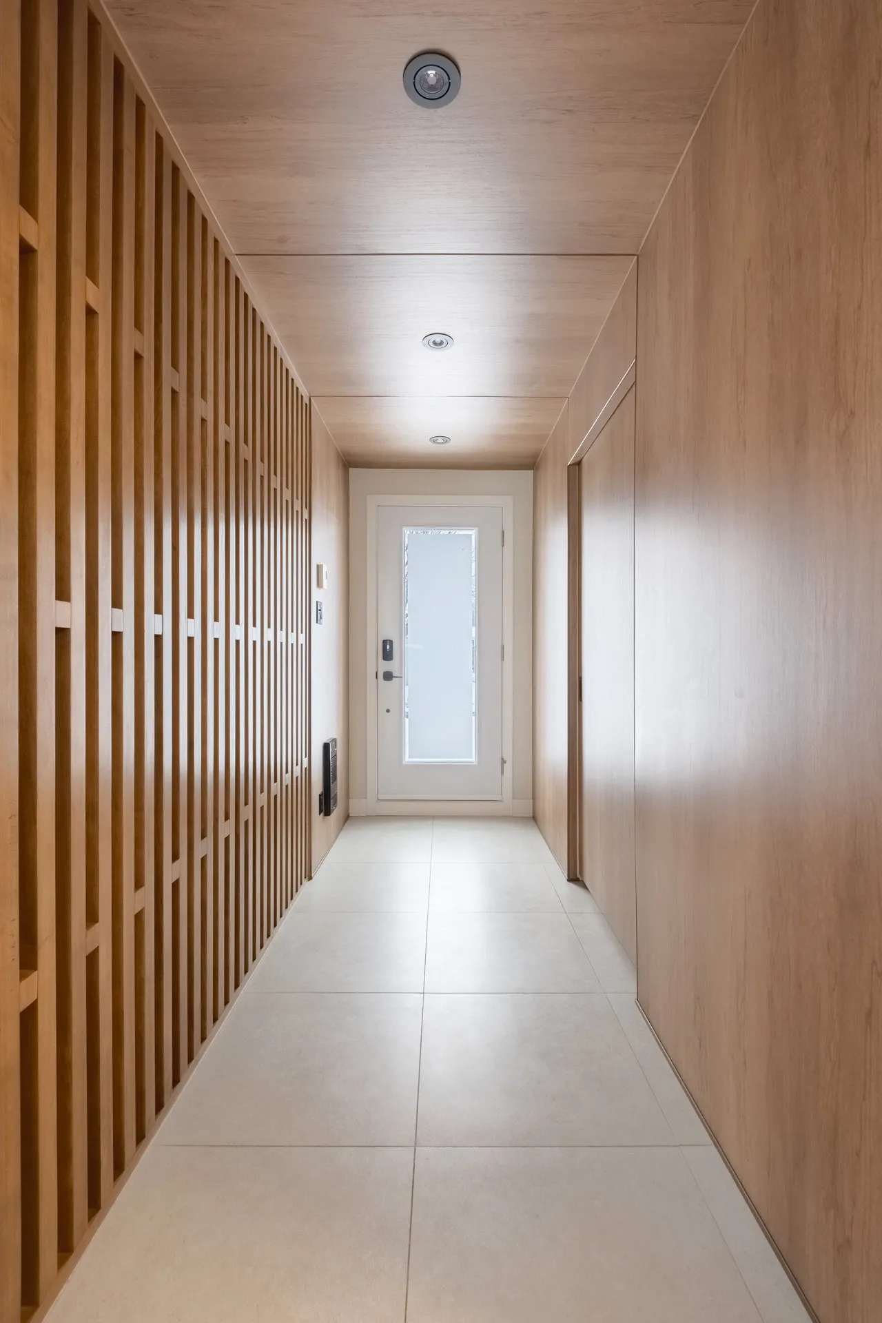 Couloir moderne avec armoires en bois clair à lattes, plancher carrelé beige, porte d'entrée blanche - Projet Léveillé Blainville