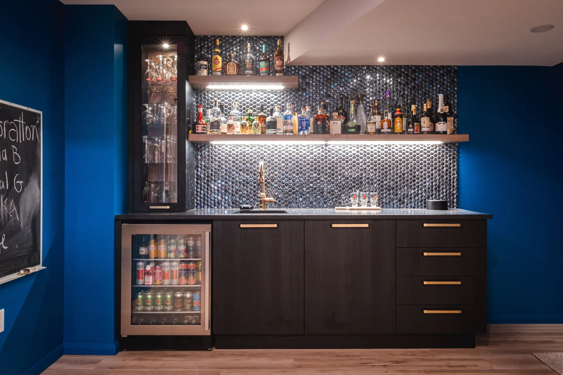Bar résidentiel bleu avec armoires noires, comptoir gris, dosseret métallisé et réfrigérateur encastré