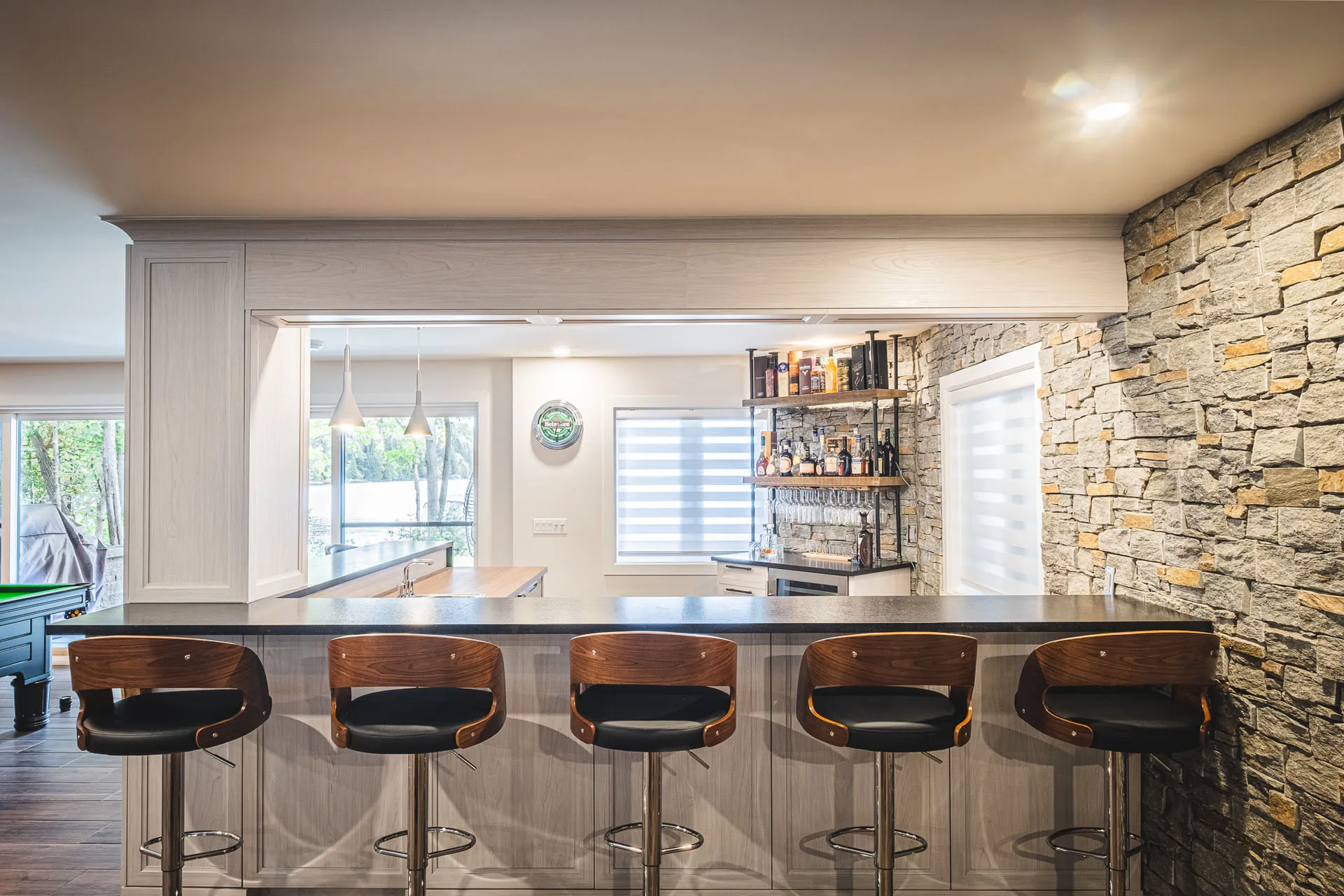 Modern kitchen with island, gray cabinets, black countertop, stone wall, wooden chairs and pendant lights