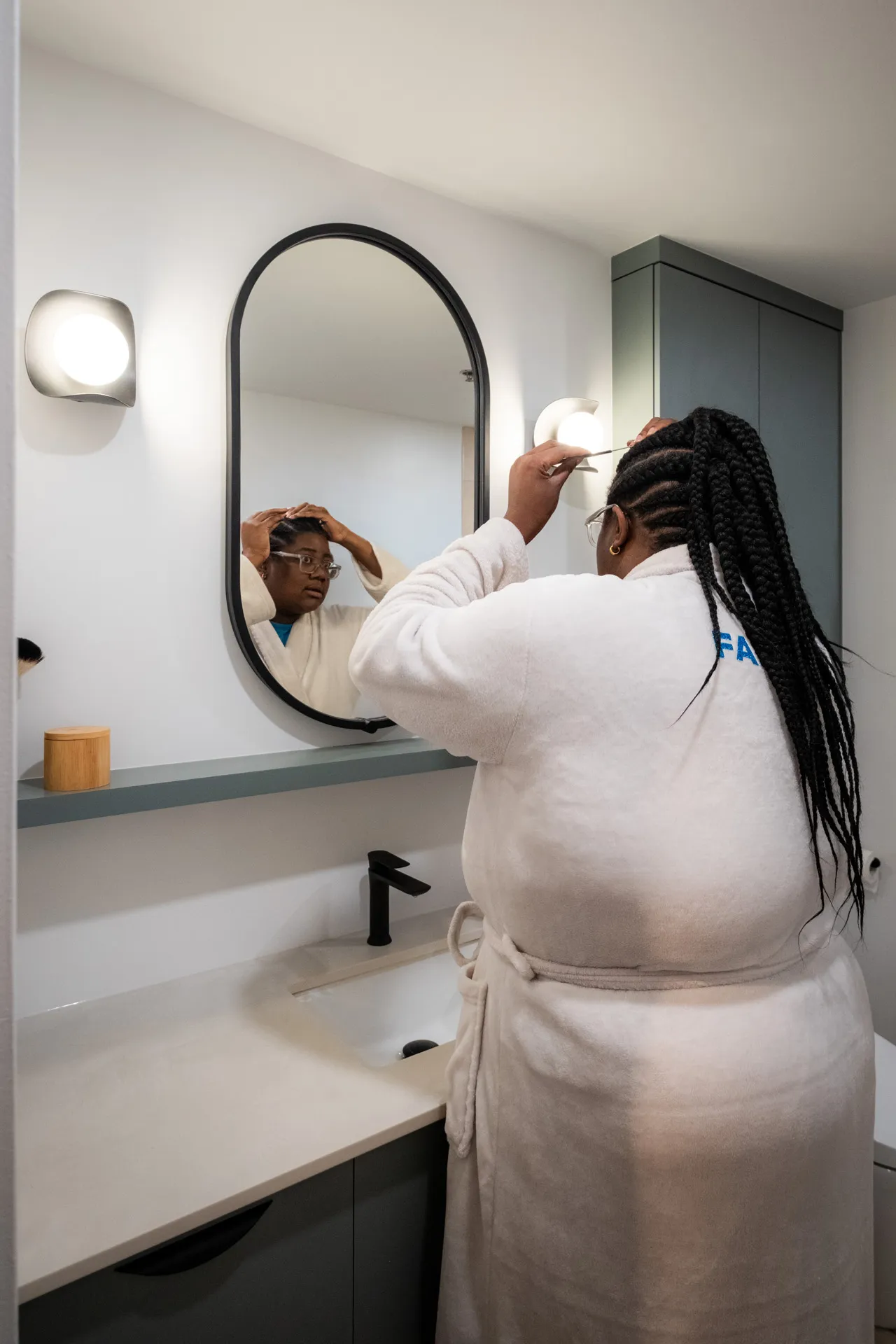 Salle de bain moderne Menuitech avec armoires grises, comptoir blanc, miroir ovale noir et luminaires muraux.