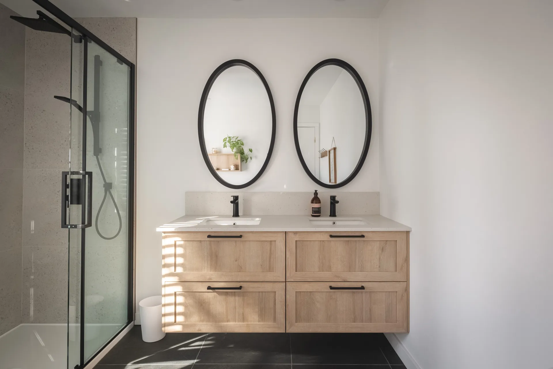 Salle de bain moderne avec vanité double en bois naturel, comptoir blanc, miroirs ovales noirs et douche vitrée