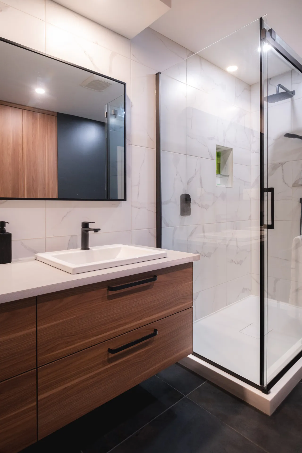 Salle de bain moderne avec vanité en bois, comptoir blanc, miroir encadré noir et douche vitrée carrelage marbre