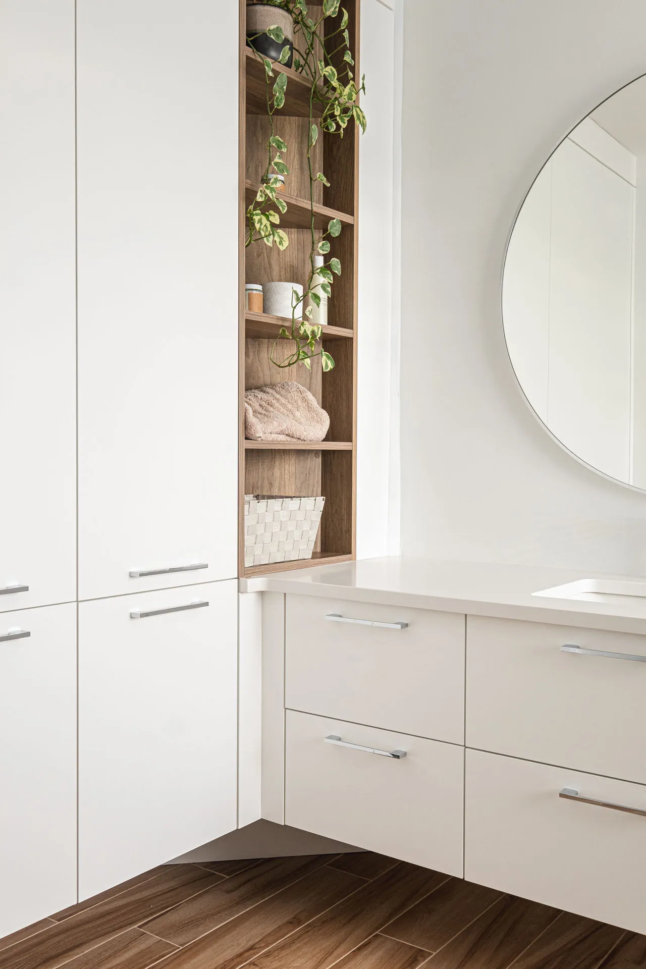 Modern bathroom with white cabinetry, wooden wall shelving, round mirror, white countertop and dark wood flooring
