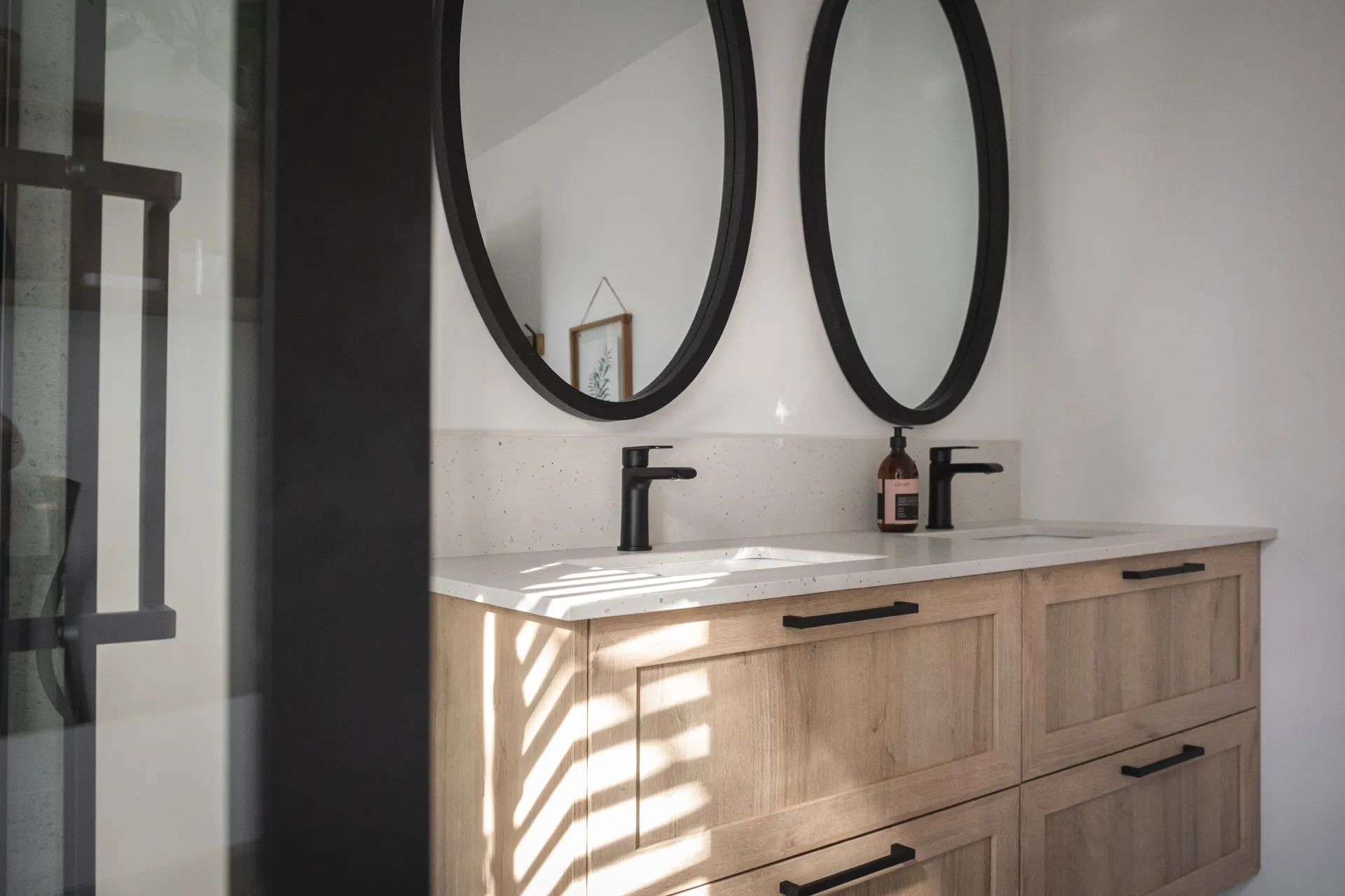 Salle de bain Diamant : vanité bois clair, comptoir blanc marbré, miroirs ovales noirs, robinetterie noire, dosseret blanc