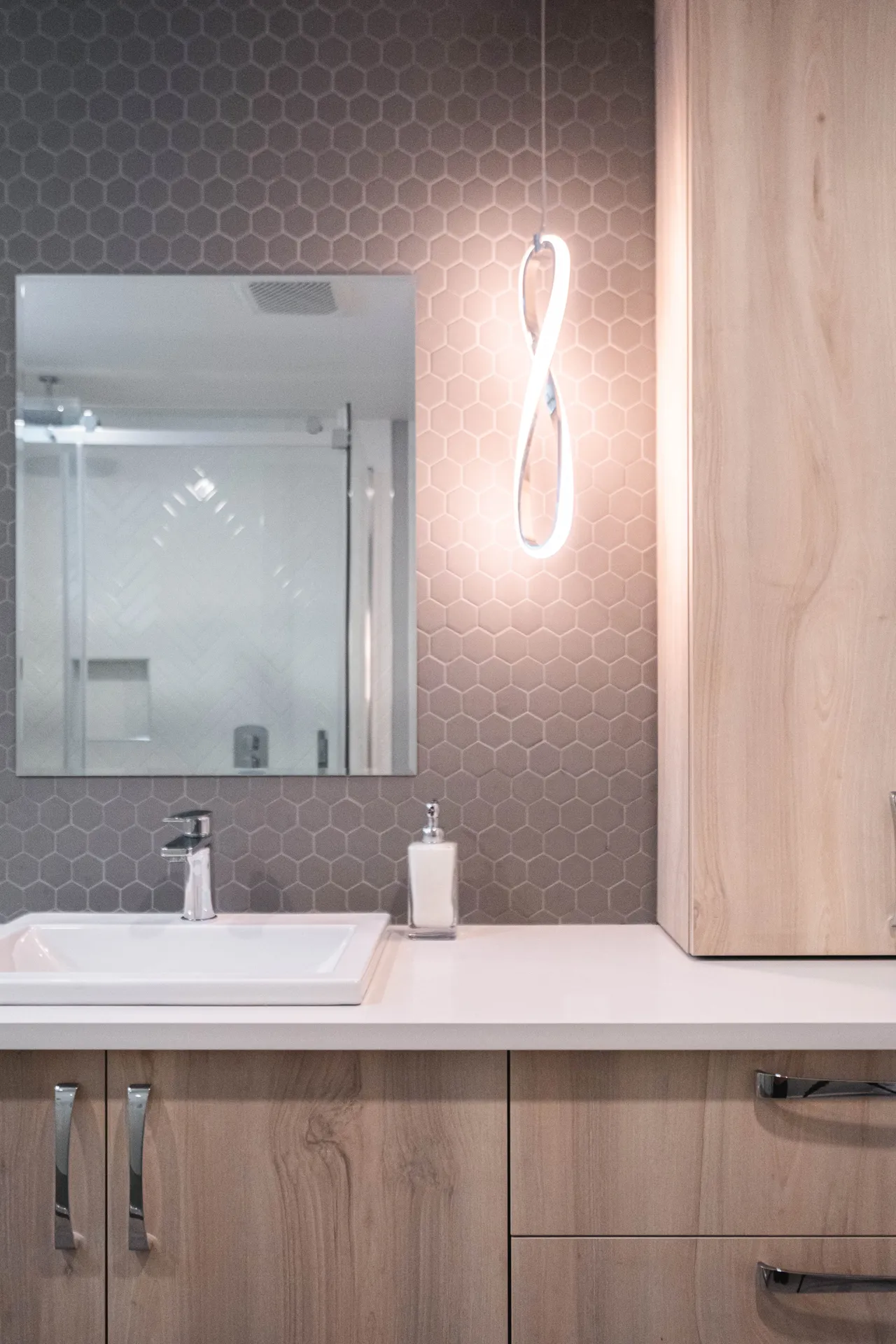 Salle de bain Menuitech avec armoires en bois clair, comptoir blanc, dosseret hexagonal gris et miroir éclairé