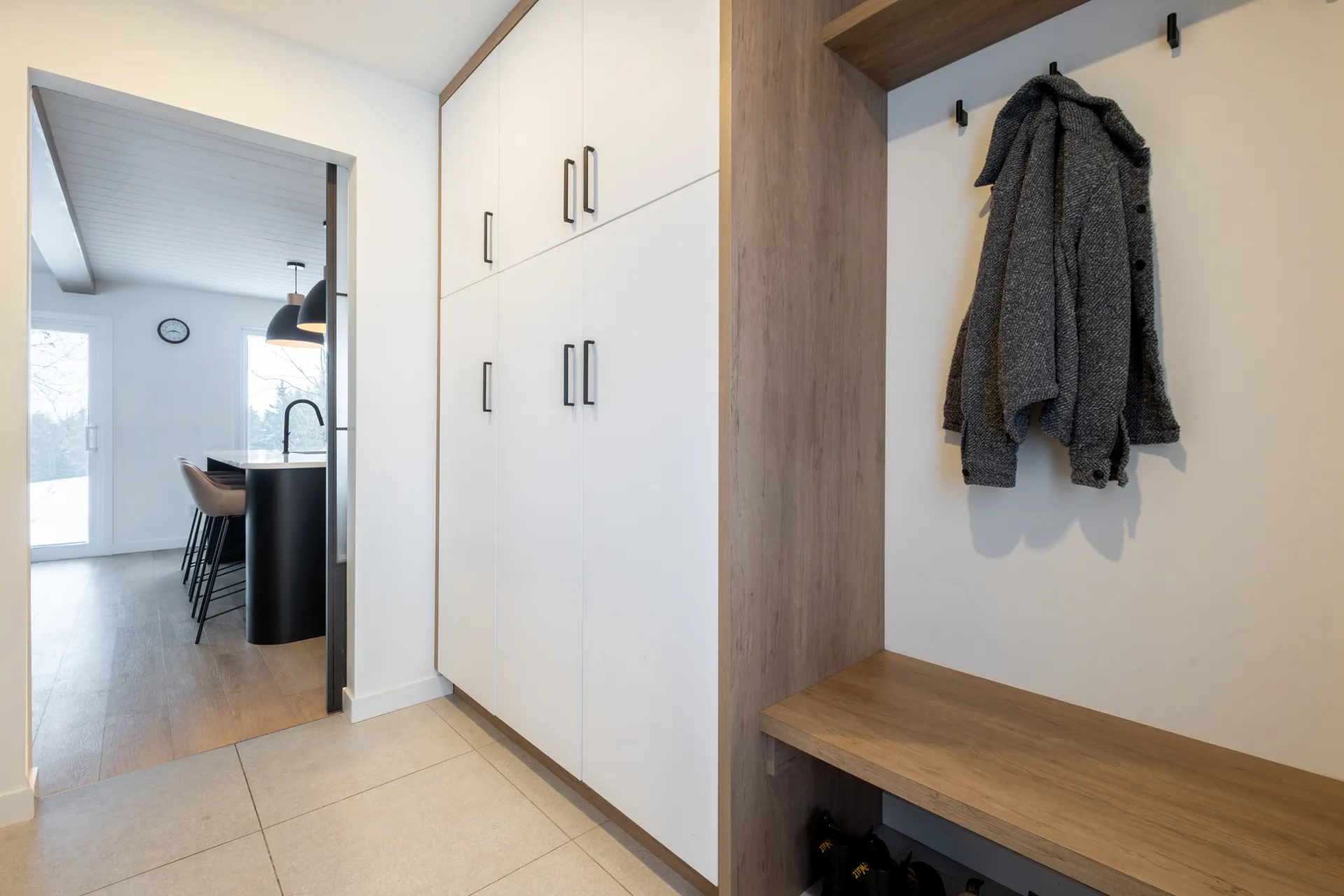 Entrée moderne avec armoires blanches sur mesure, banc bois et crochets muraux, cuisine visible au fond