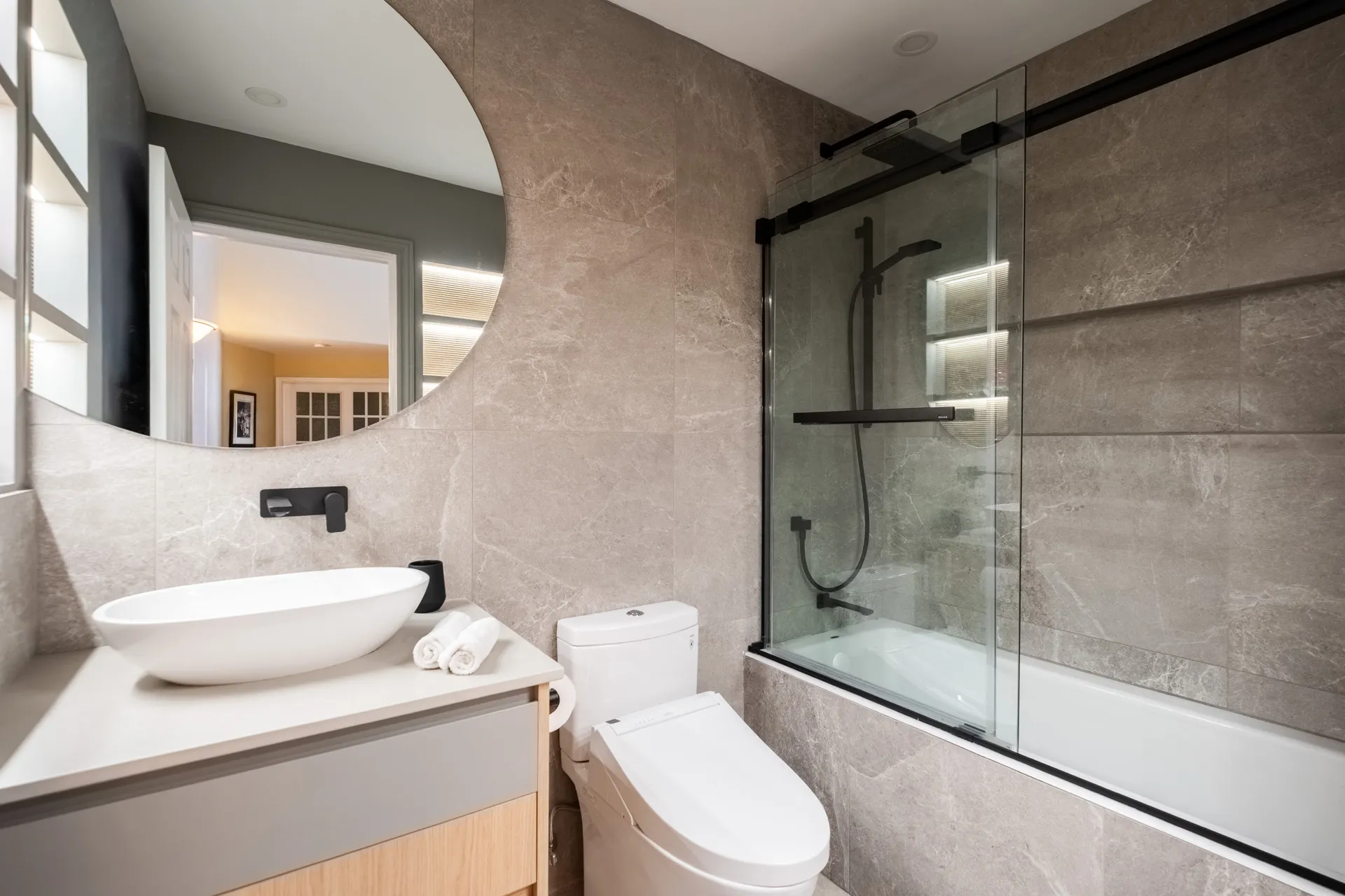 Salle de bain moderne Menuitech : vanité bois naturel, comptoir blanc, lavabo rond blanc, miroir courbe éclairé, douche vitrée noire.