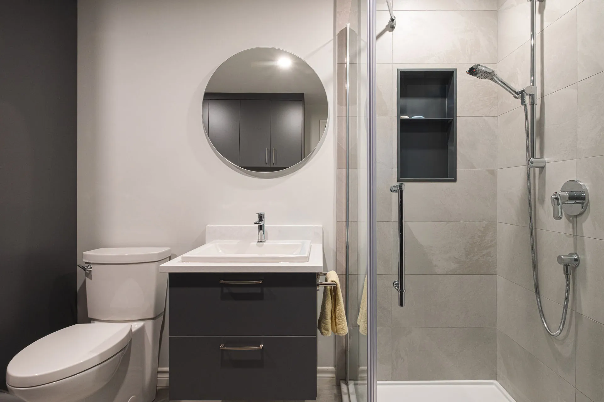 Modern bathroom with black cabinets, white countertop, round mirror and glass shower enclosure - Menuitech renovation Boisbriand