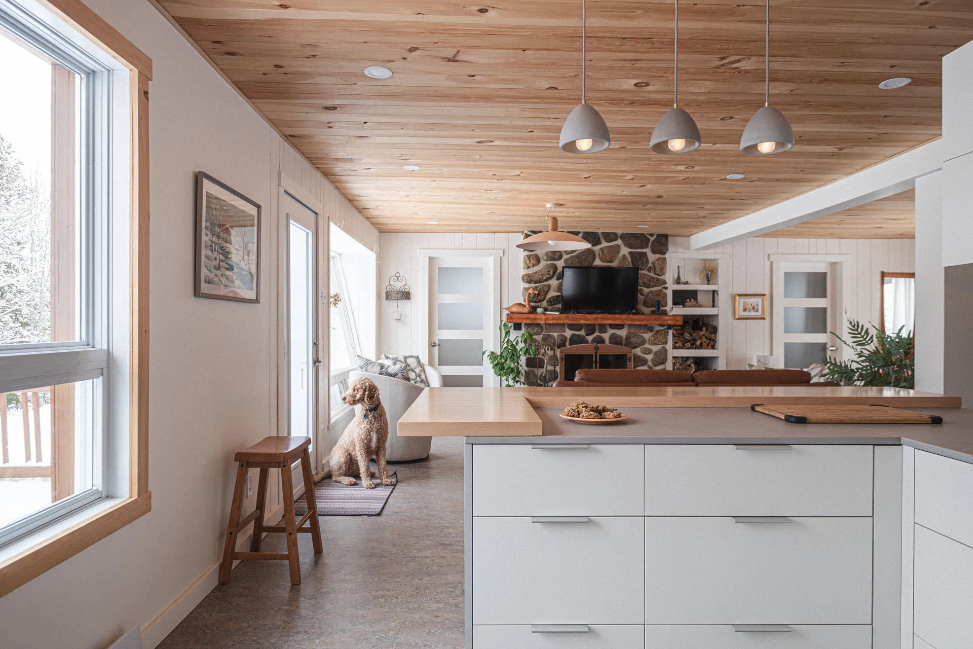 Open kitchen with white cabinets, wood countertop, island, stone fireplace, wood ceiling, gray pendant lights