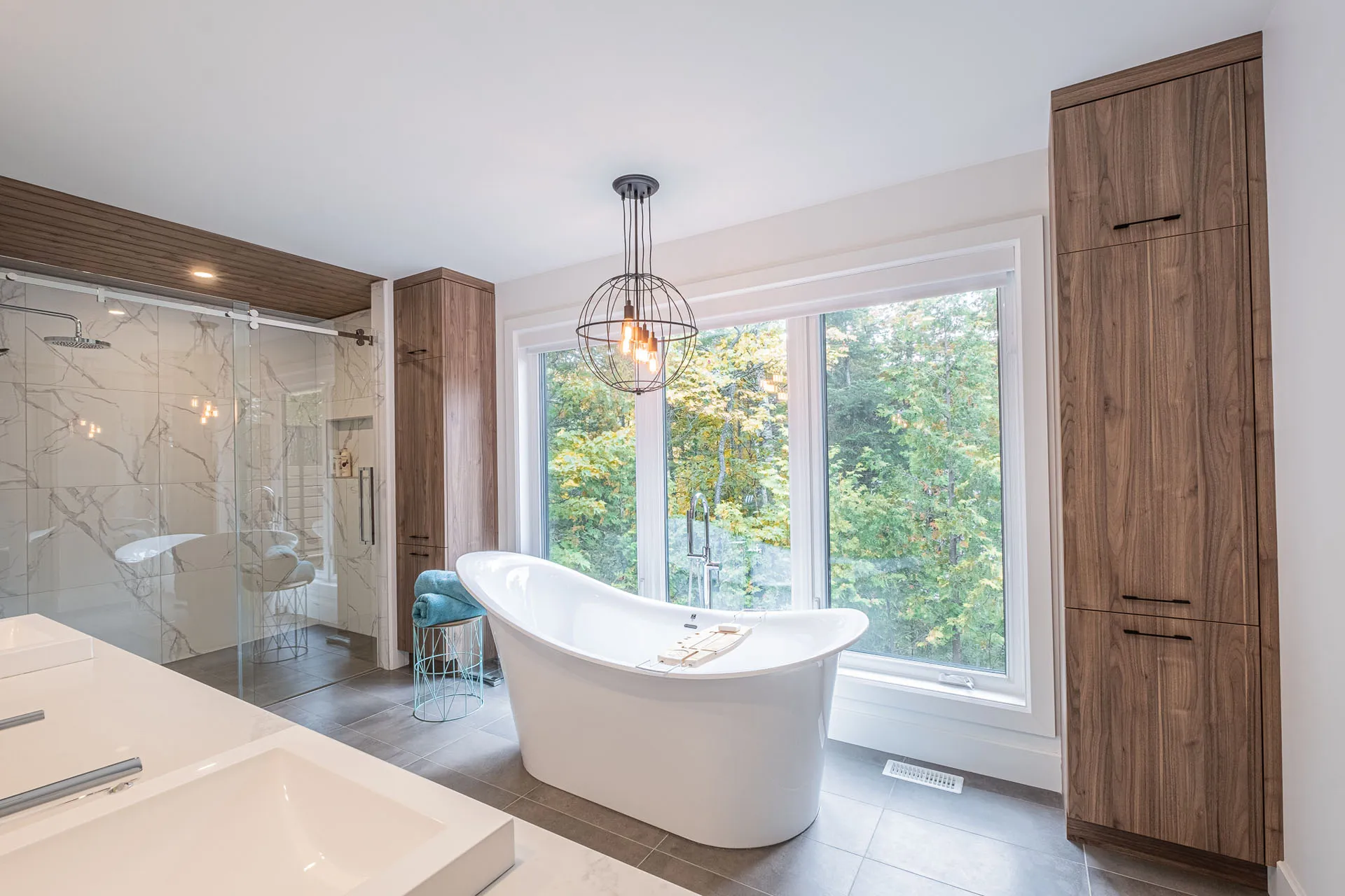 Modern bathroom with walnut wood cabinets, white freestanding tub, white countertop, glass shower enclosure and large windows