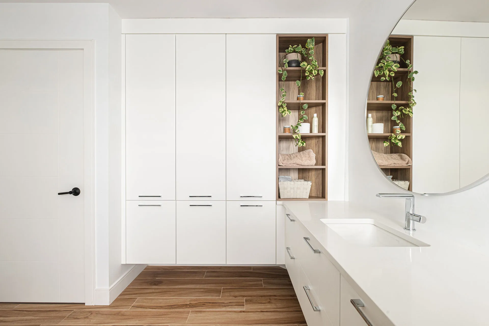 Modern bathroom with custom white cabinetry, white countertop, round mirror, wooden shelves with plants and storage solutions