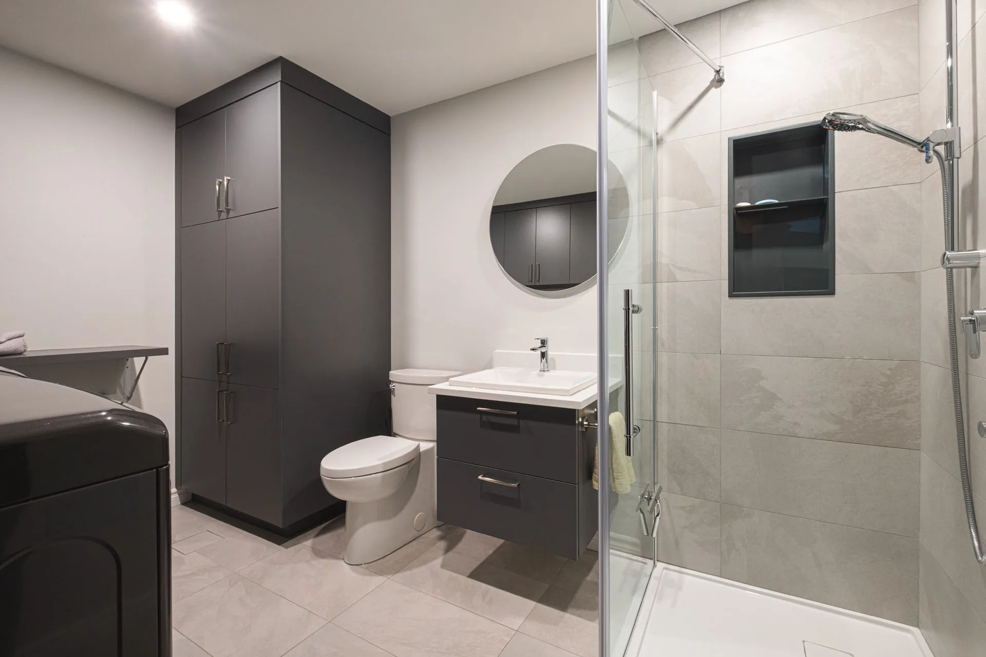 Modern bathroom with black cabinetry, white countertop, round mirror and glass shower enclosure - Landry-D'amours project Boisbriand