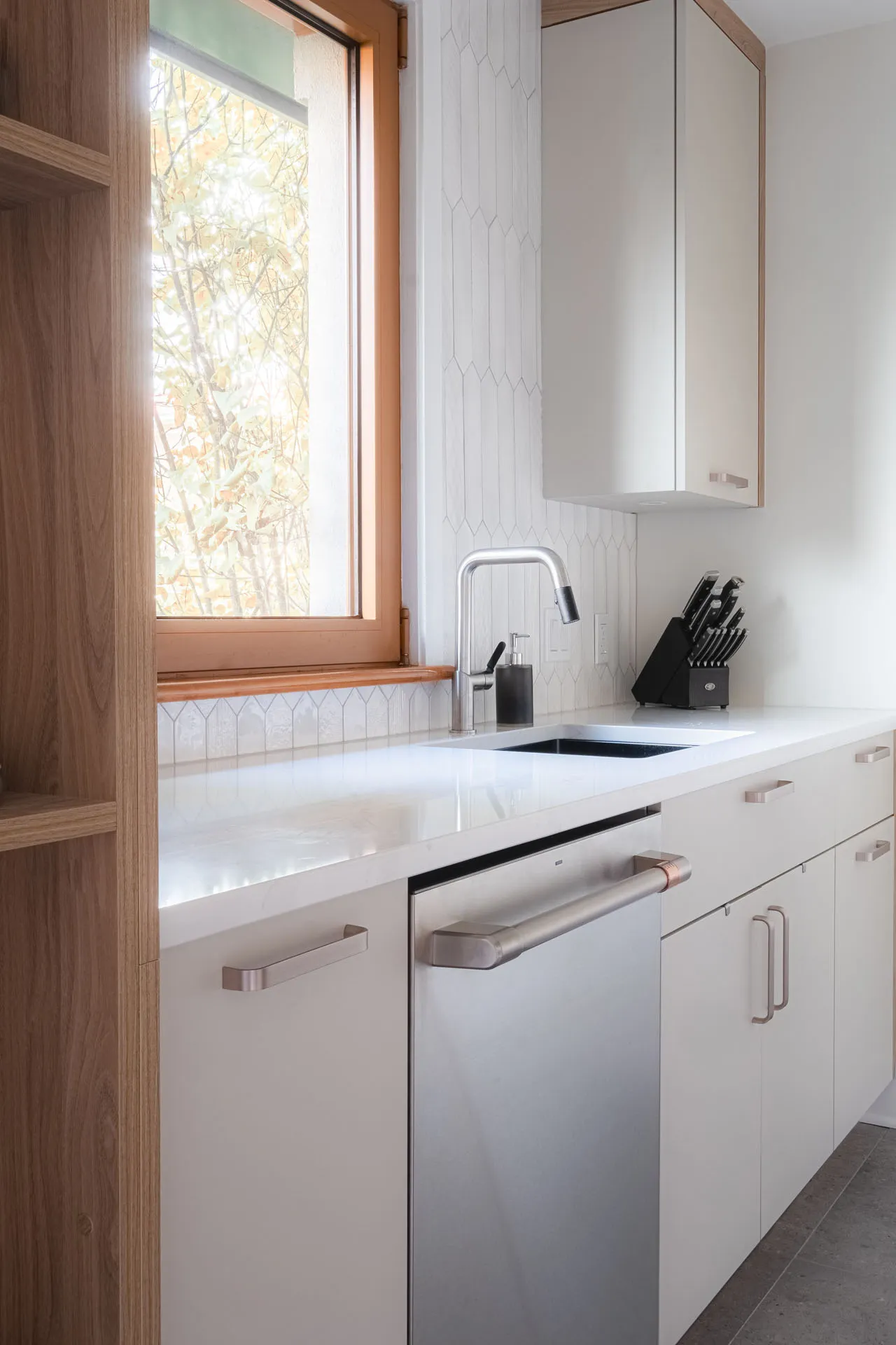 Minimalist white kitchen with white countertop, custom cabinetry, geometric backsplash and wood window frame.