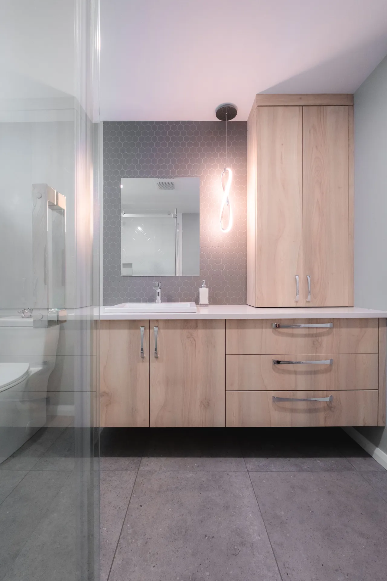 Salle de bain moderne avec vanité en bois clair, comptoir blanc, dosseret hexagonal gris et luminaire néon rose
