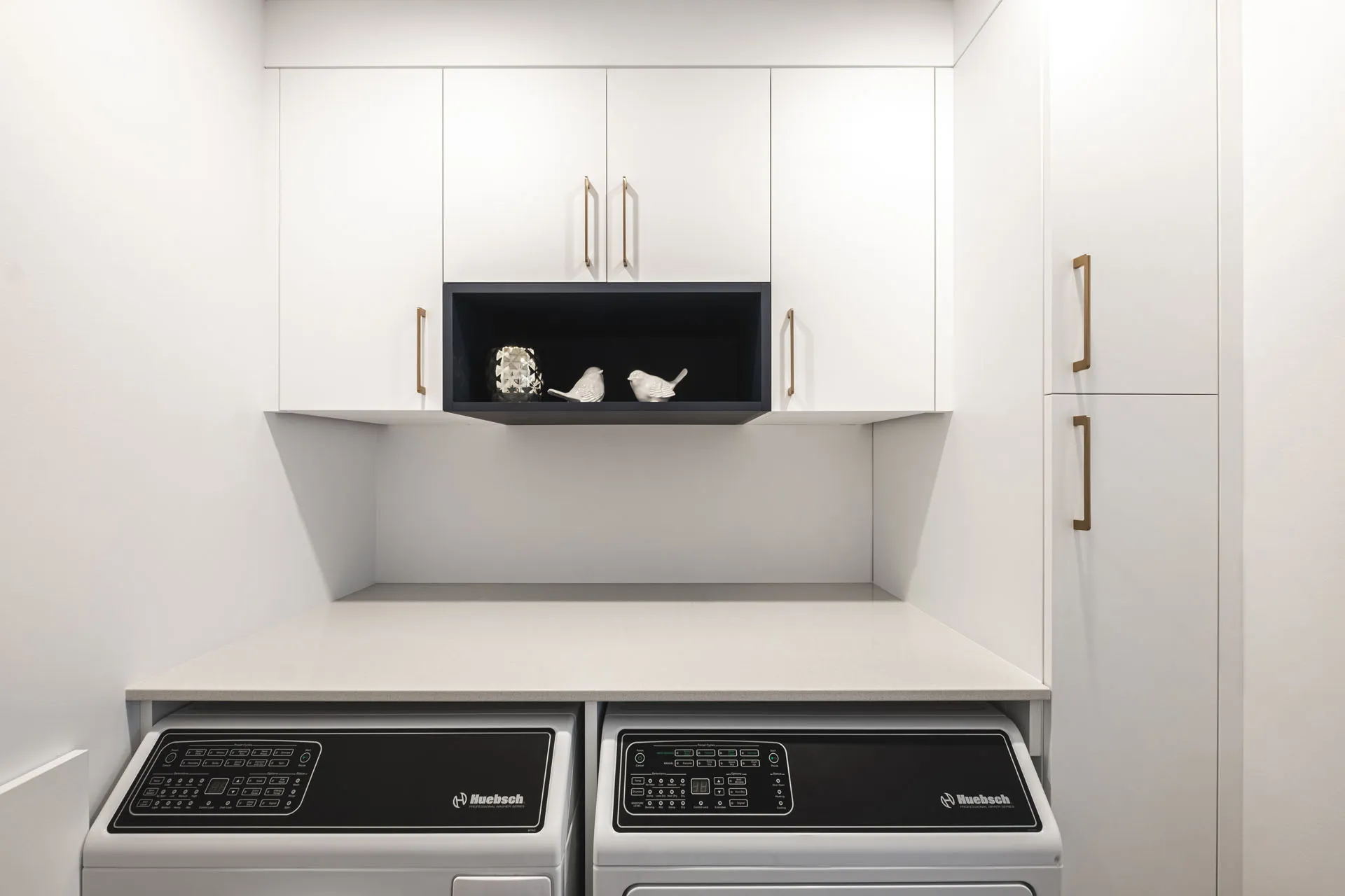 Modern Menuitech laundry room: white cabinets, light grey countertop, black niche with decor, two Huebsch washers