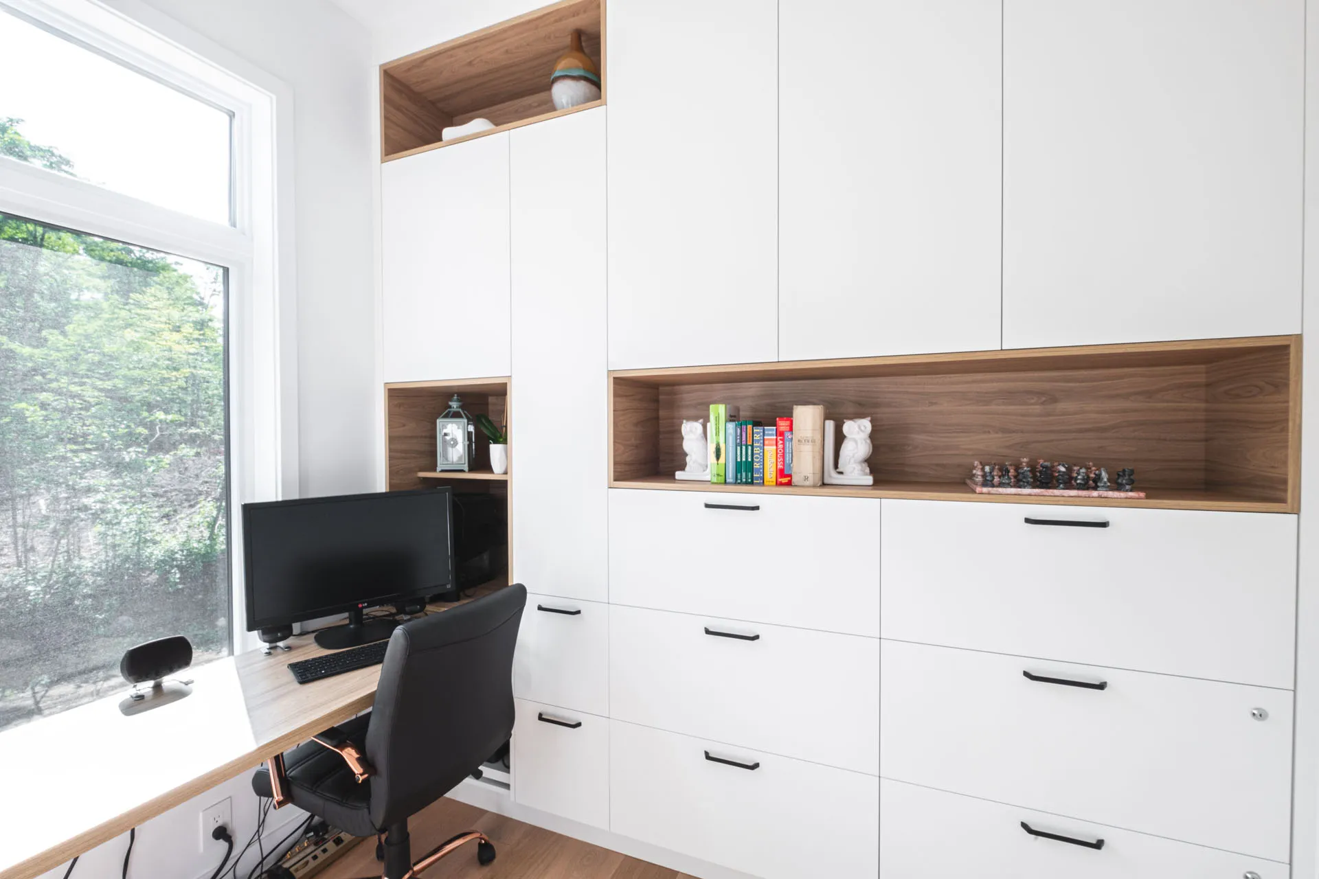 White and wood office with custom cabinetry, wood countertop, open shelving, window, black chair, monitor