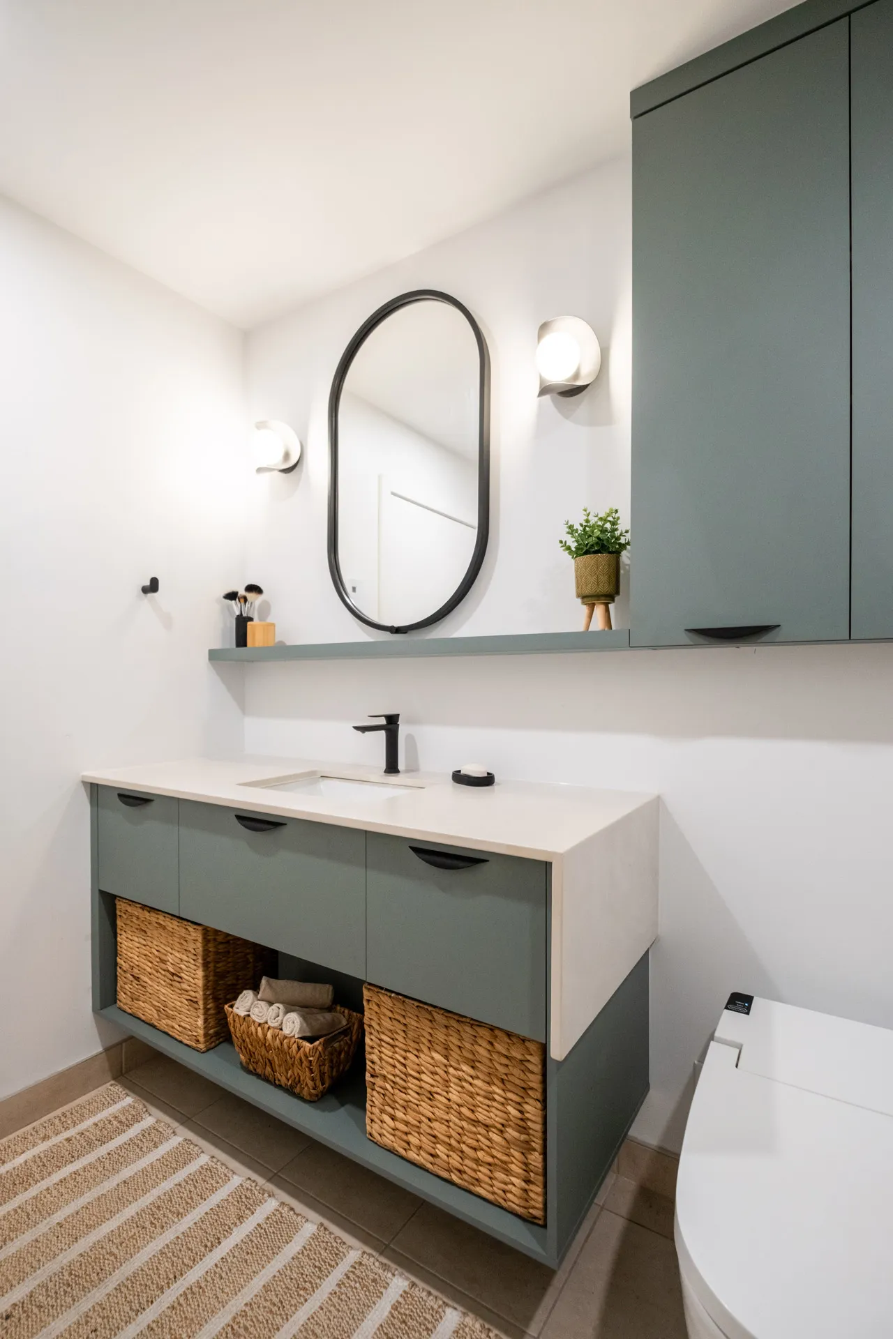 Salle de bain moderne avec vanité vert sauge, comptoir blanc, miroir ovale noir et rangements en osier