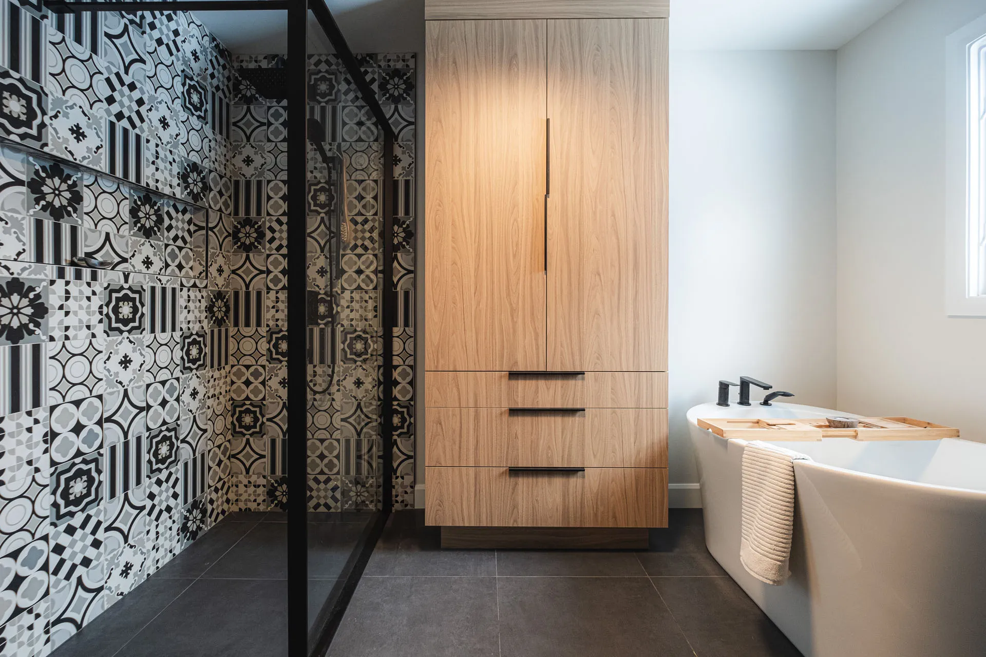 Robitaille bathroom with natural wood cabinets, black and white tile backsplash, white bathtub and grey countertop