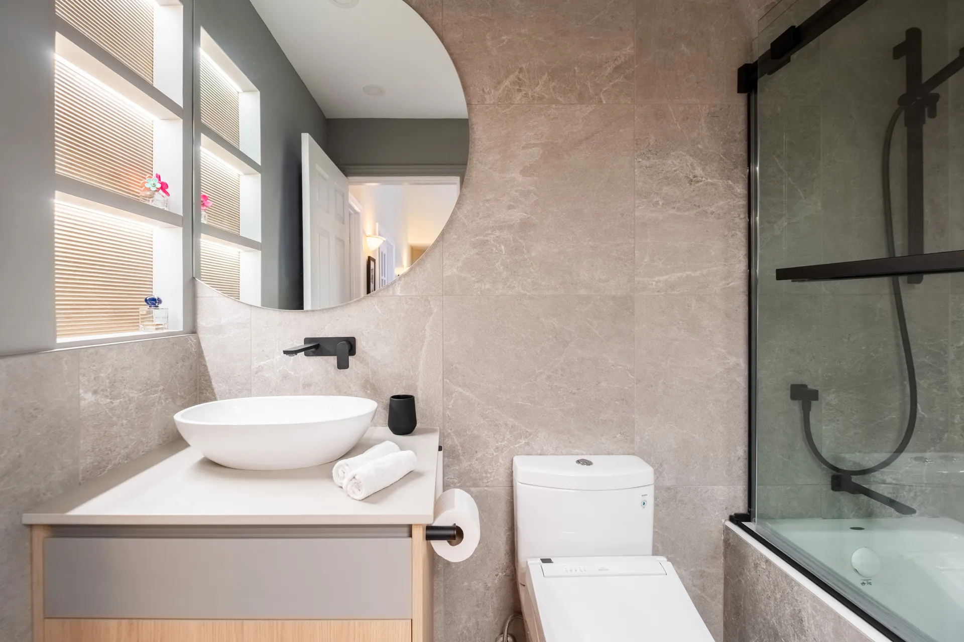 Salle de bain moderne Garraud Rosemère: armoires blanches et bois, comptoir blanc, carrelage beige, miroir rond, douche vitrée