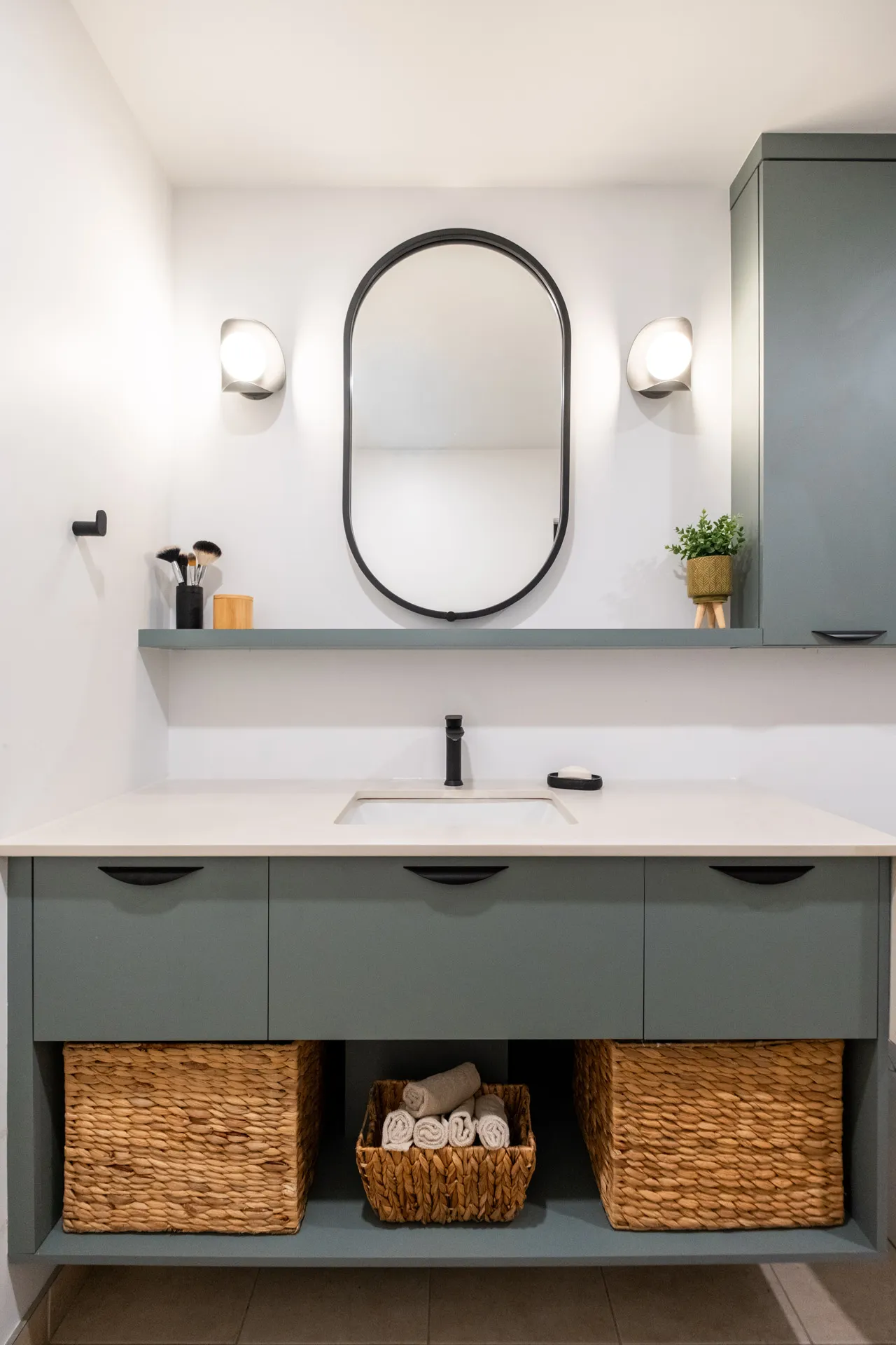 Salle de bain Fabiola : vanité grise avec comptoir blanc, miroir ovale noir, appliques murales et paniers de rangement.
