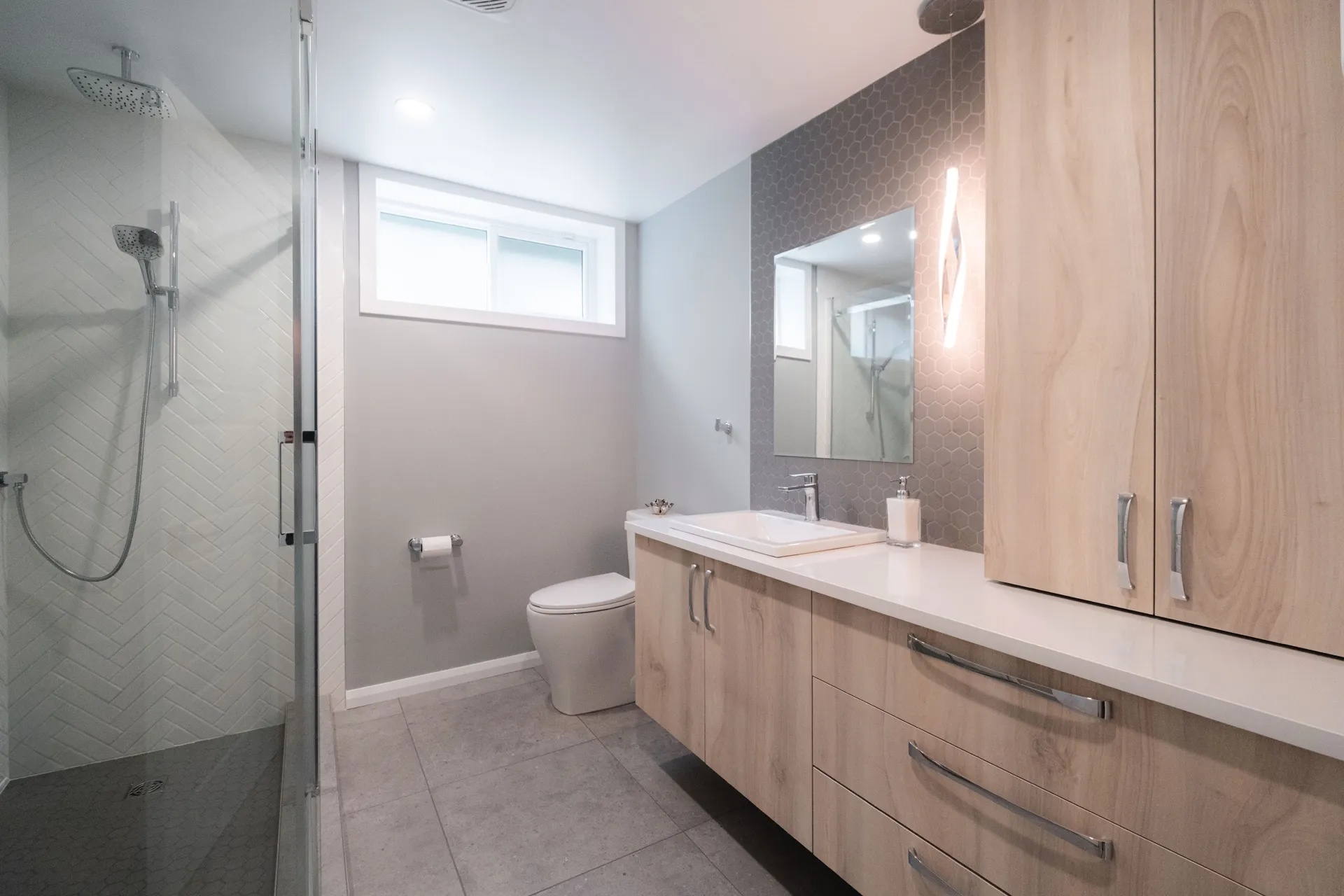 Salle de bain moderne avec armoires en bois clair, comptoir blanc, miroir éclairé et douche vitrée