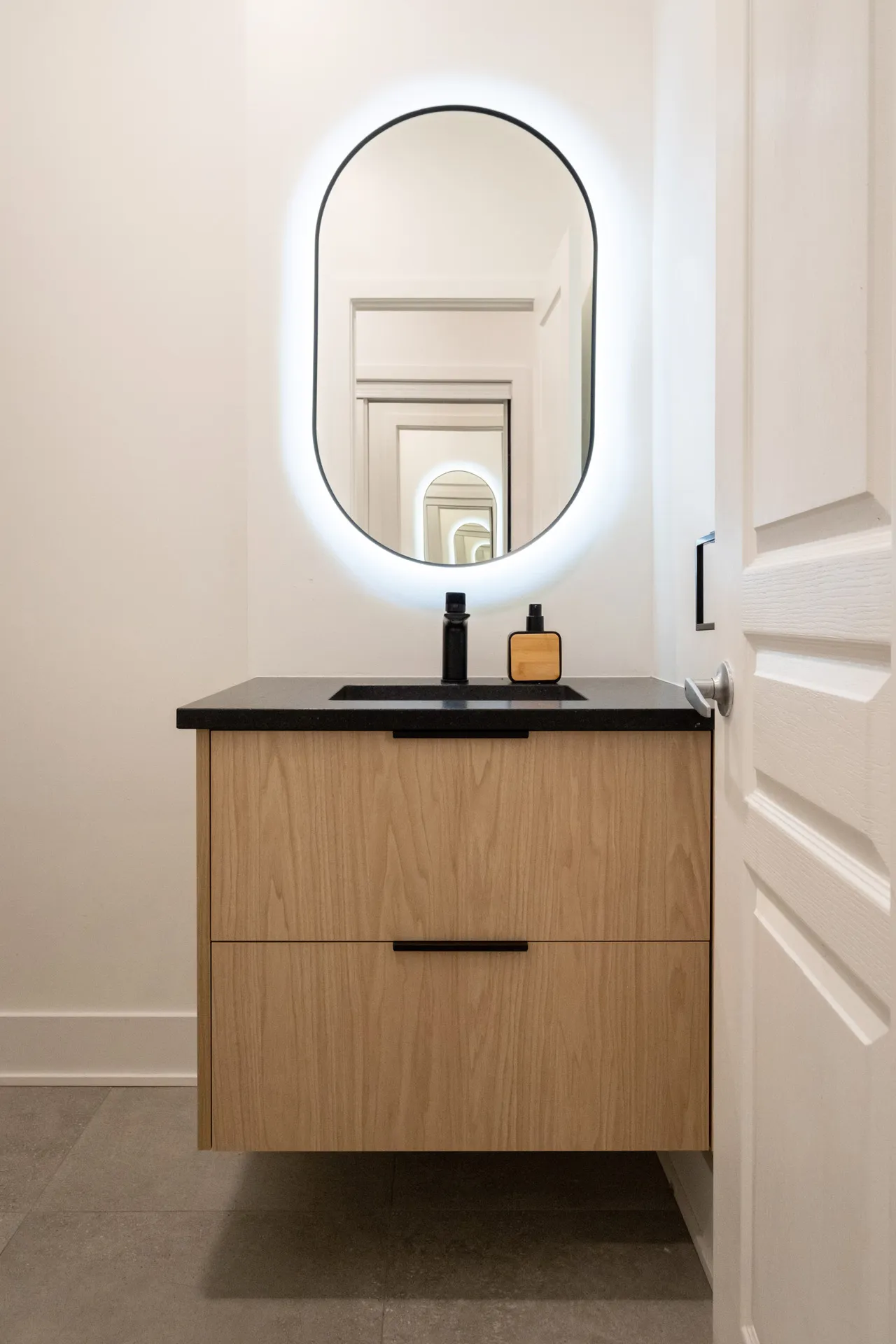 Salle de bain Pelletier : vanité en bois clair, comptoir noir, miroir ovale lumineux LED, robinetterie noire