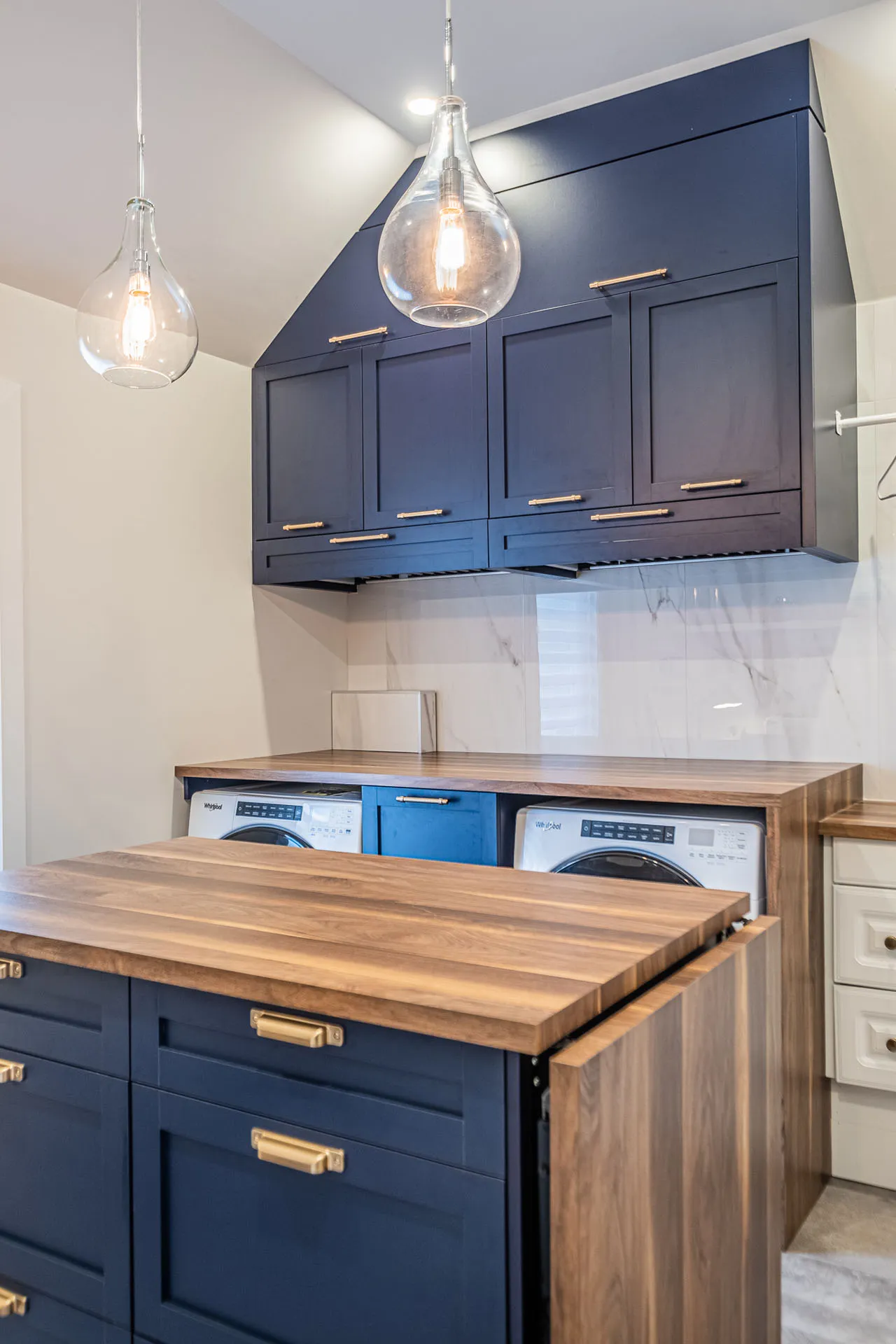 Simard laundry room: navy blue Menuitech cabinets, wood countertop, island with marble backsplash, glass pendant lights, built-in washers