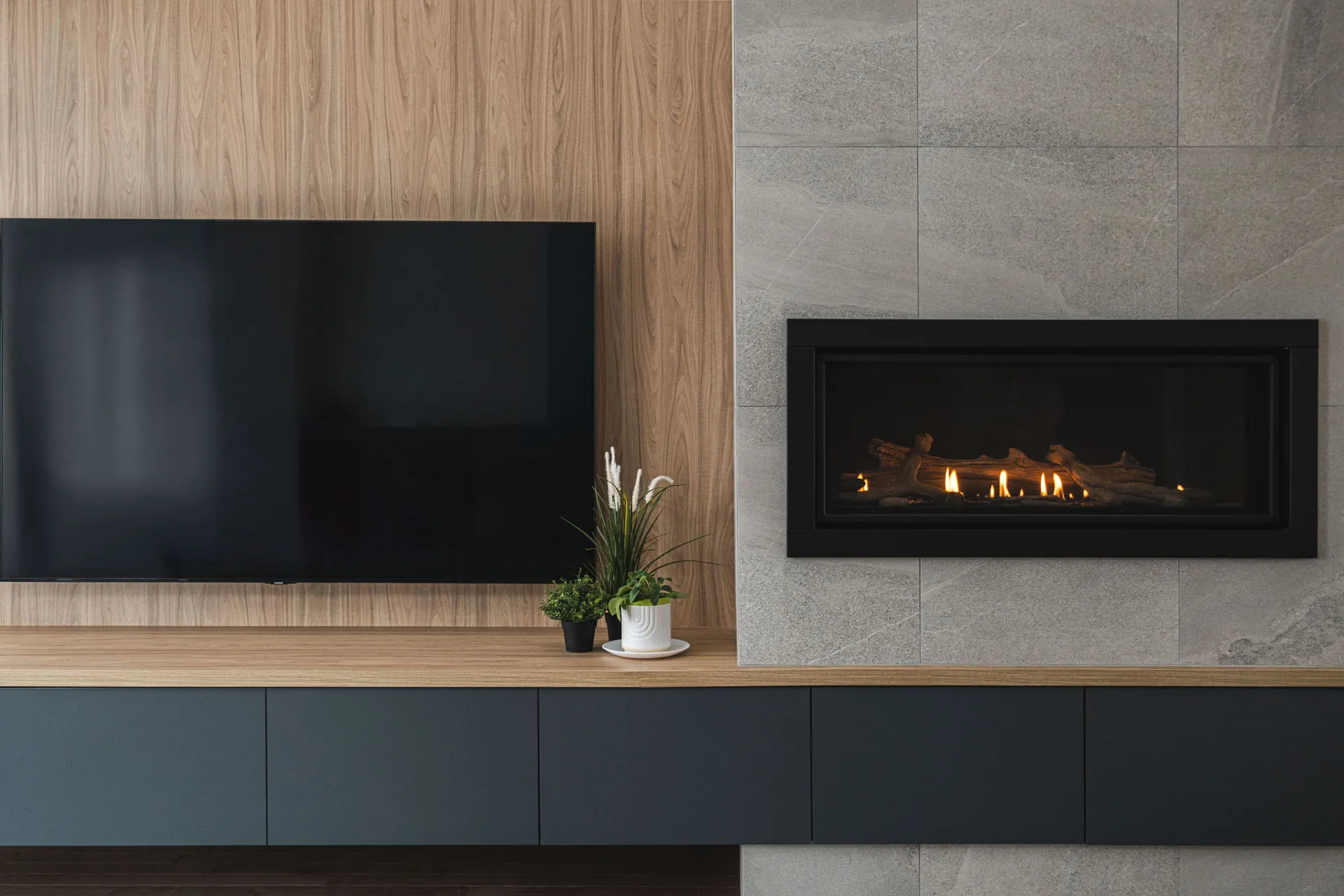 Modern Boisbriand kitchen: grey-blue cabinets, light wood countertop, grey backsplash with integrated fireplace, wall-mounted TV