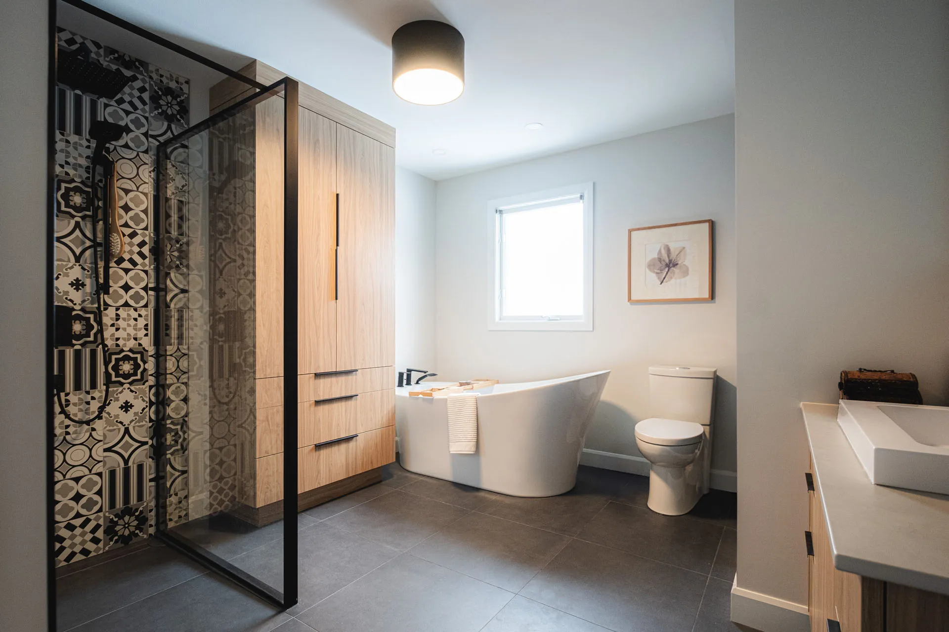 Modern bathroom with light wood cabinetry, white soaking tub, toilet, grey countertop and geometric patterned shower enclosure