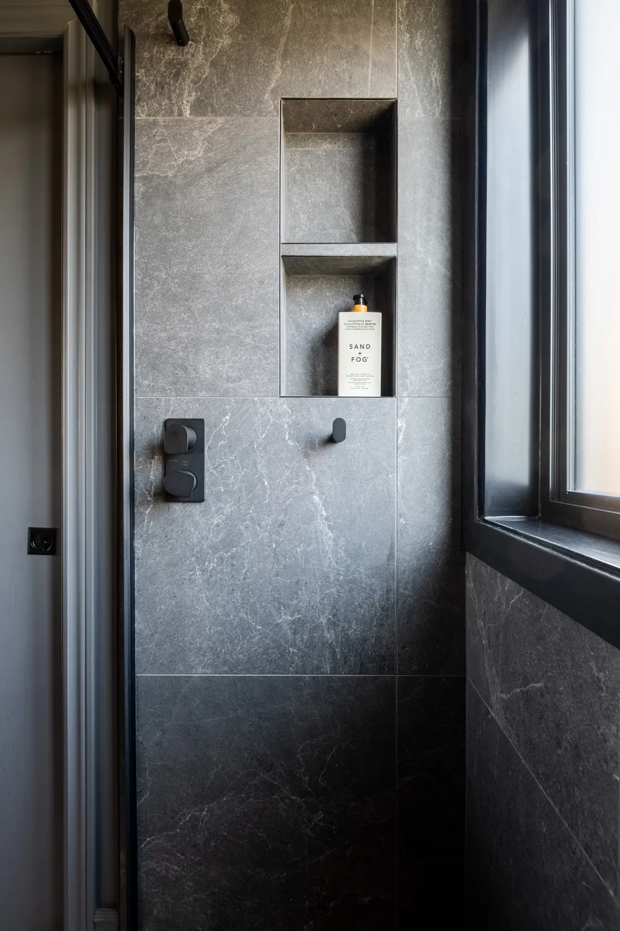 Douche contemporaine avec carrelage gris marbre, niches intégrées, robinetterie noire et fenêtre - Projet Garraud Rosemère