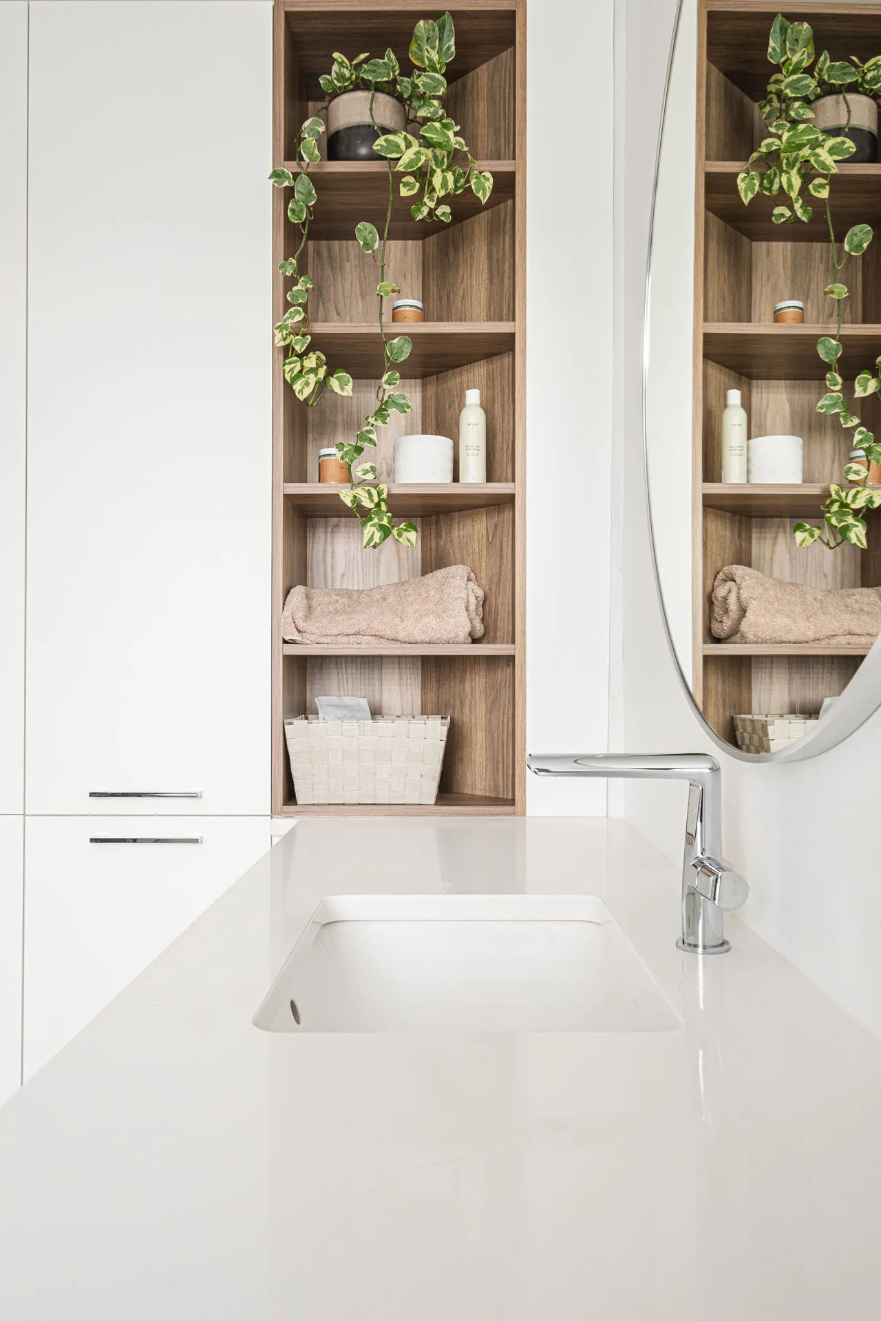 Modern bathroom with white cabinetry, white countertop, oval mirror and wooden wall shelves with plants