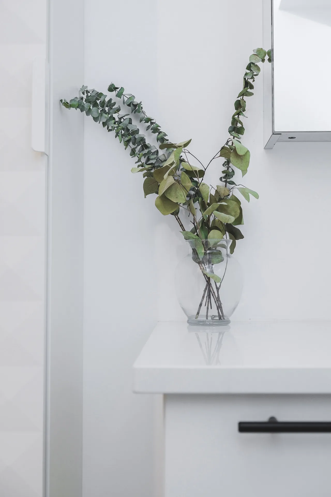 Minimalist white bathroom with white countertop, sleek cabinetry and eucalyptus floral arrangement in glass vase