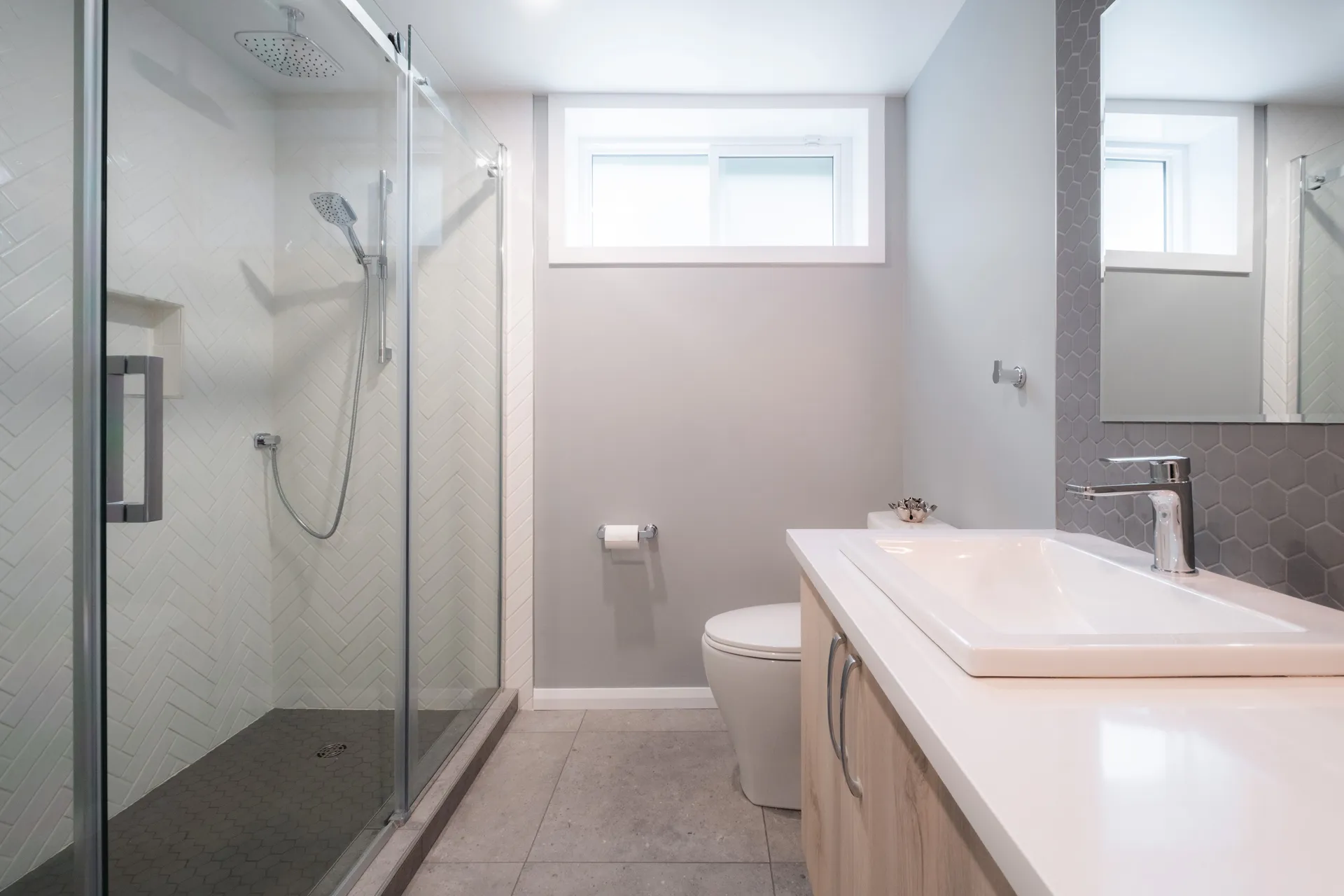 Salle de bain moderne avec vanité bois clair, comptoir blanc, dosseret gris chevron et douche vitrée carrelage blanc