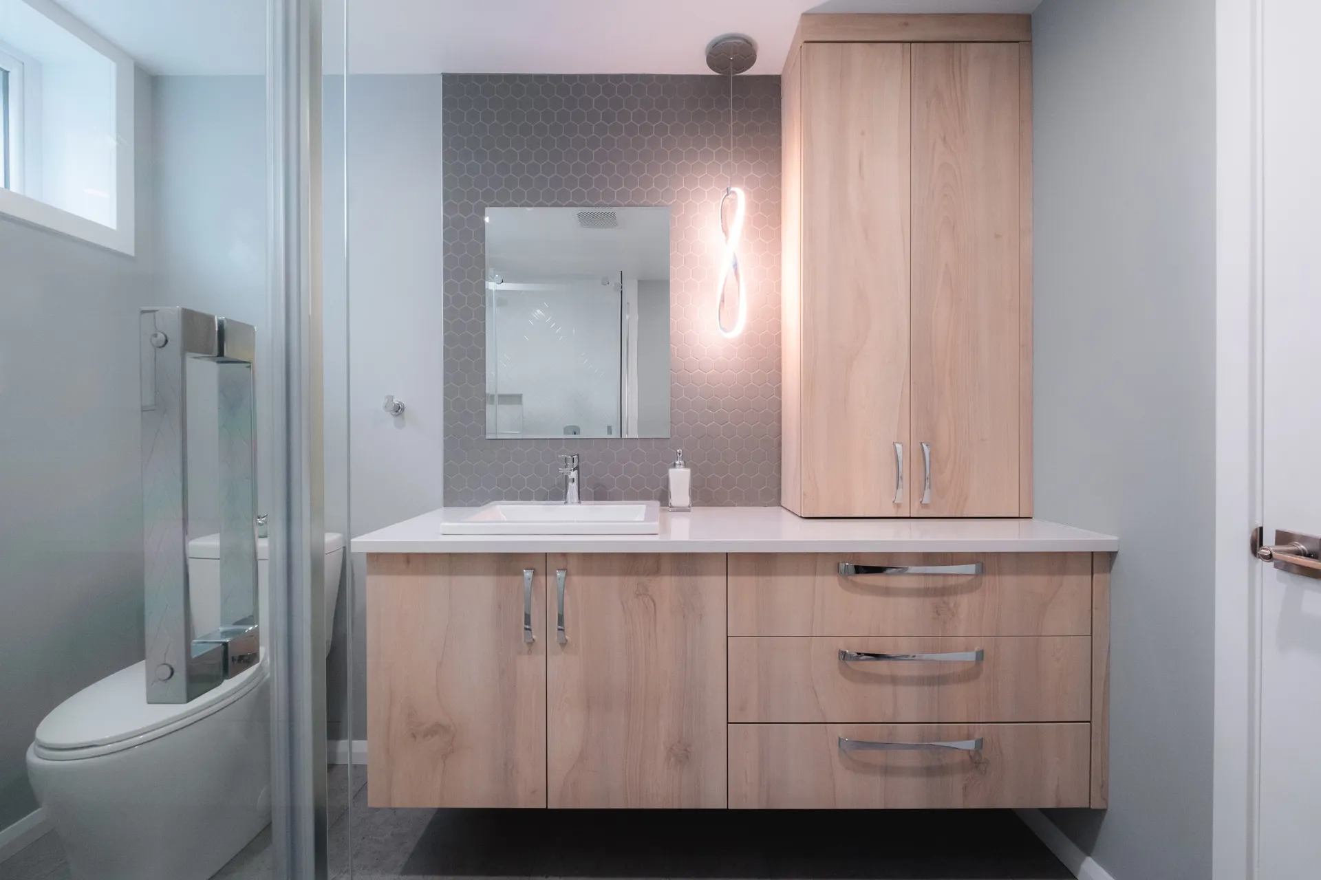 Salle de bain moderne avec vanité en bois clair, comptoir blanc, miroir encadré et dosseret hexagonal gris