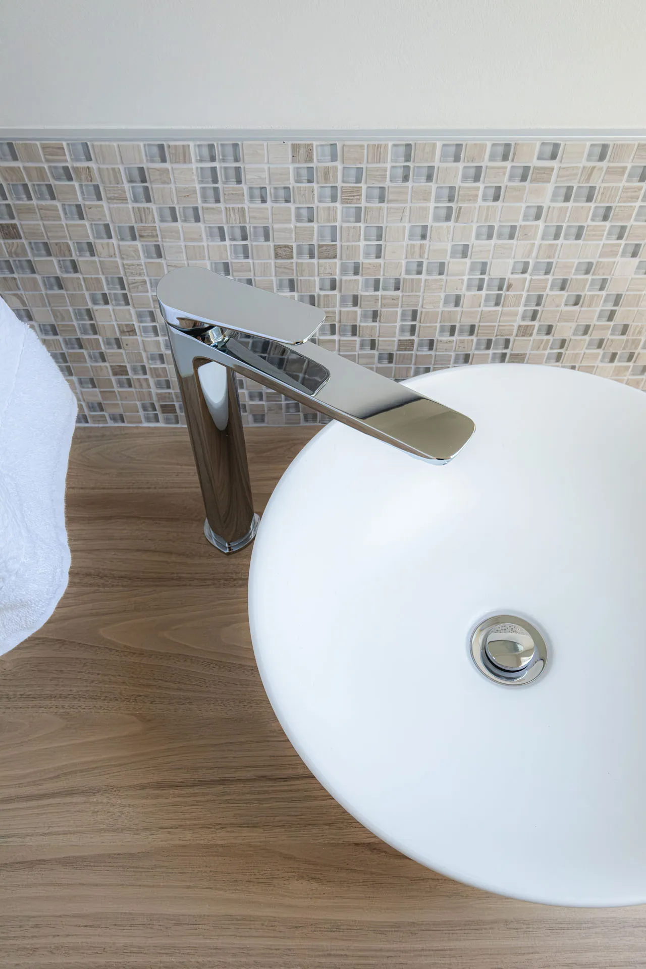 Salle de bain moderne avec lavabo rond blanc, robinetterie chromée, dosseret en mosaïque beige et sol en bois.