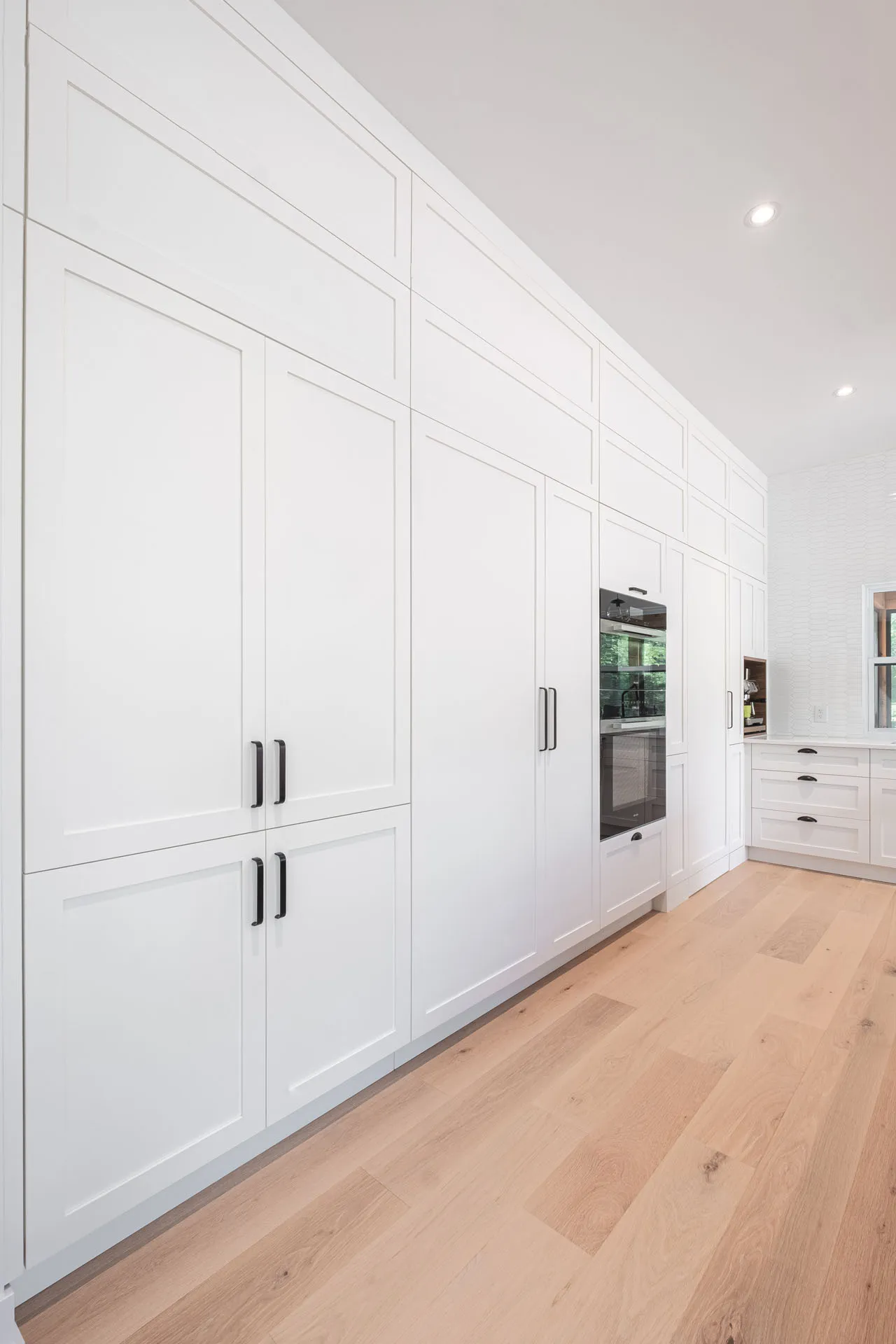 Modern white Menuitech kitchen with shaker cabinets, built-in oven, light wood flooring and recessed lighting