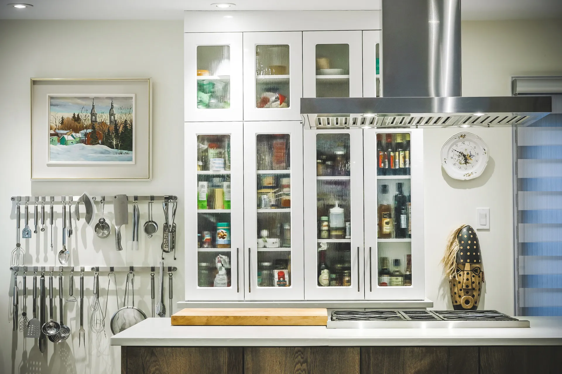 White kitchen with glass-front cabinets, white countertop, wooden island, wall-mounted utensil storage, tiled backsplash, stainless hood