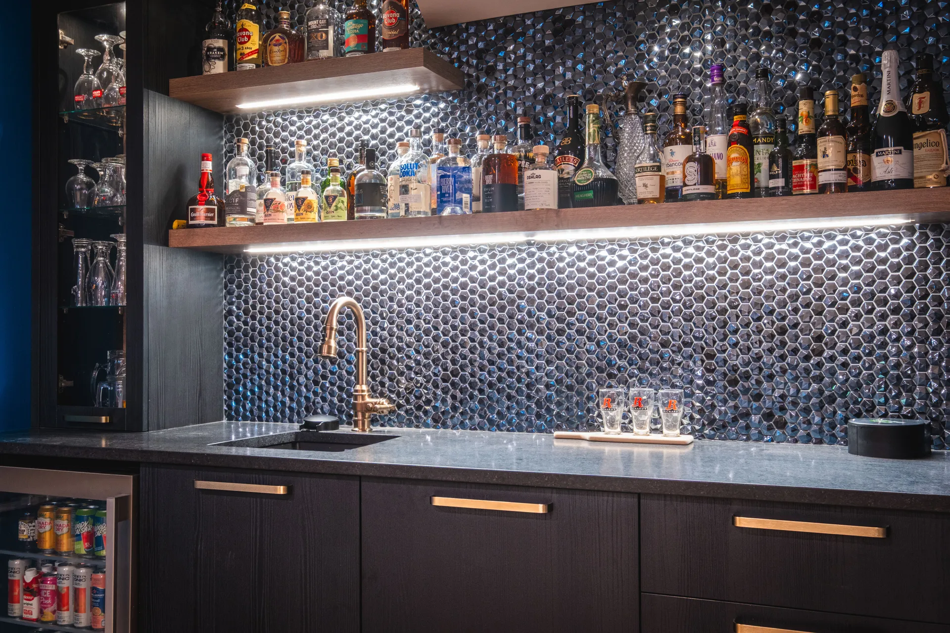 Bar résidentiel Menuitech avec armoires noires, comptoir gris, dosseret hexagonal métallisé et étagères en bois éclairées