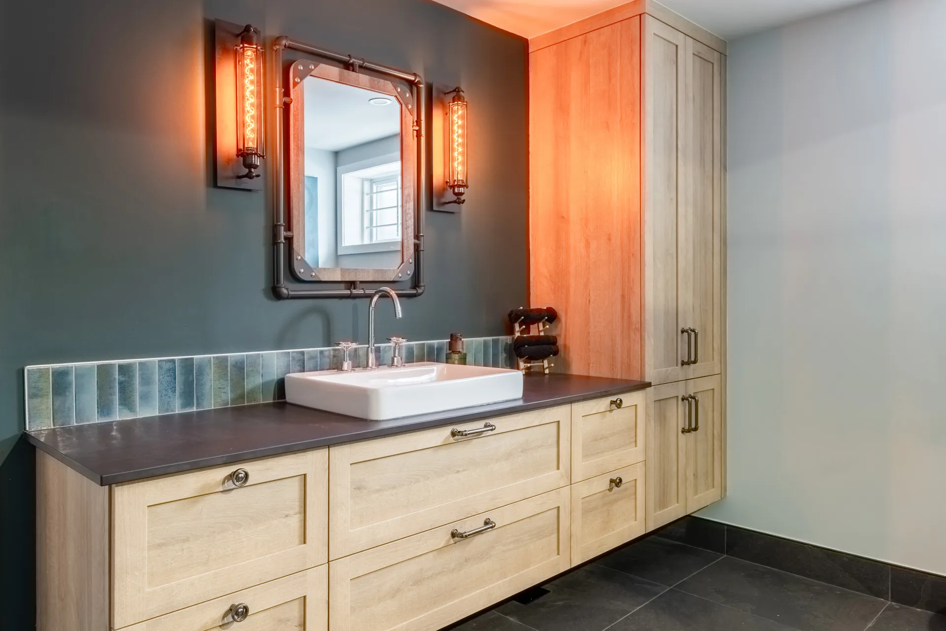 Lemay bathroom with light wood vanity, dark grey countertop, industrial mirror and copper wall sconces