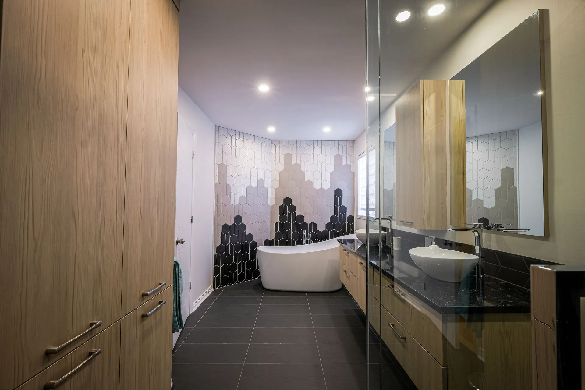 Modern Landry bathroom: light wood cabinets, black countertop, white freestanding tub, black and white hexagonal backsplash