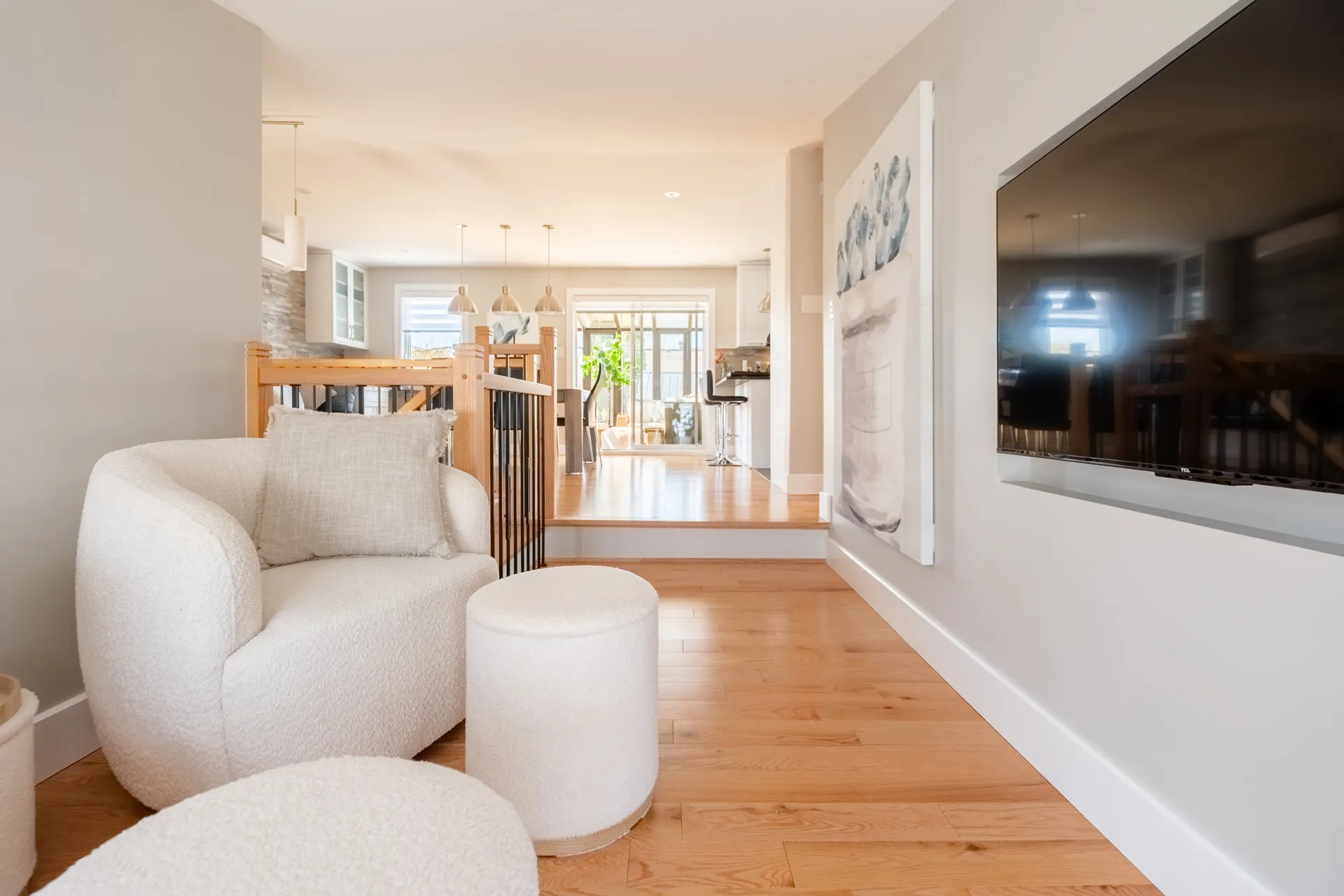 Salon moderne avec fauteuil beige, ottoman blanc et téléviseur mural, ouvert sur cuisine aux armoires bois naturel