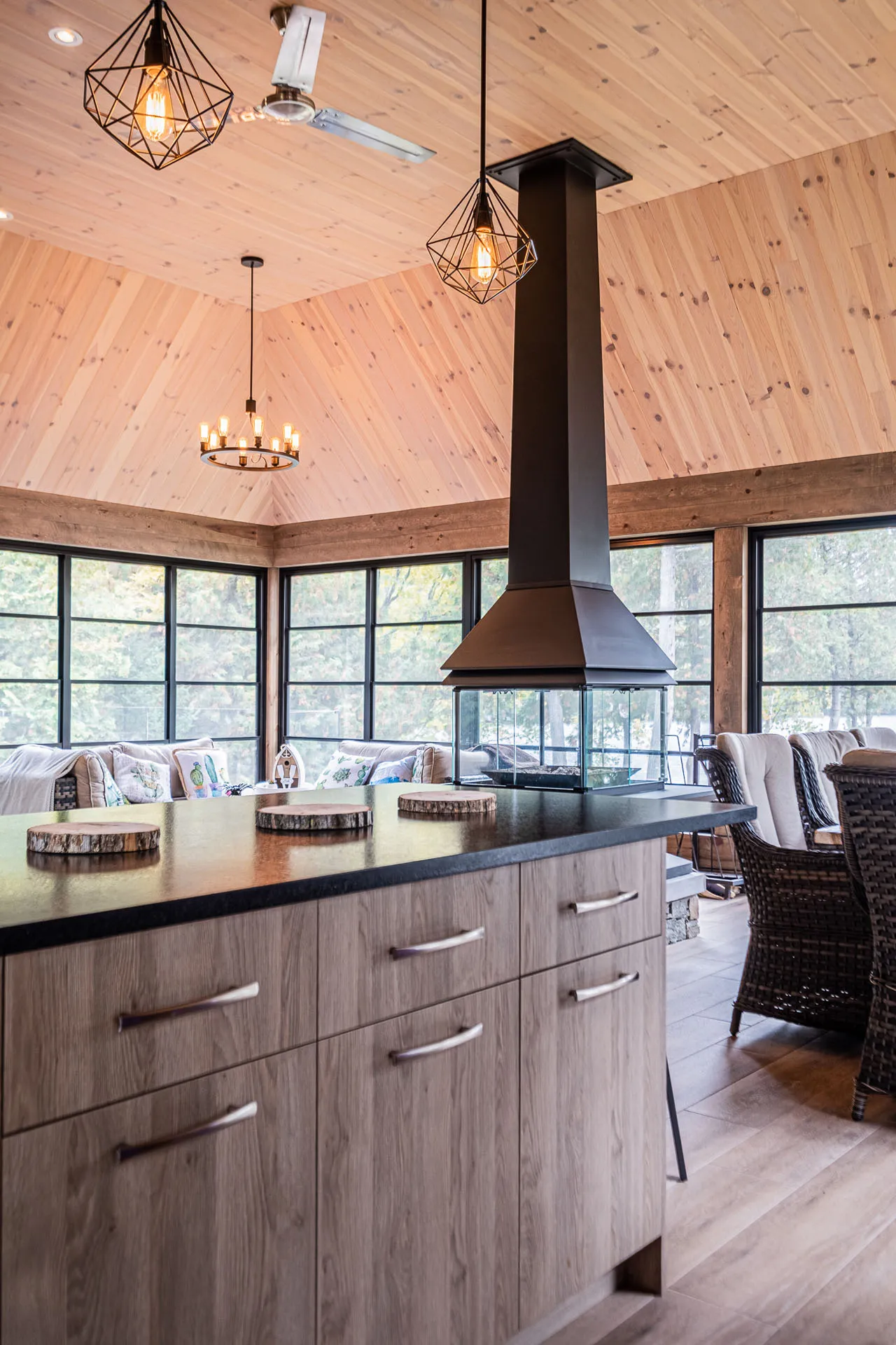 Open kitchen with island, light wood cabinets, black countertop, central fireplace, cathedral ceiling, geometric pendant lights