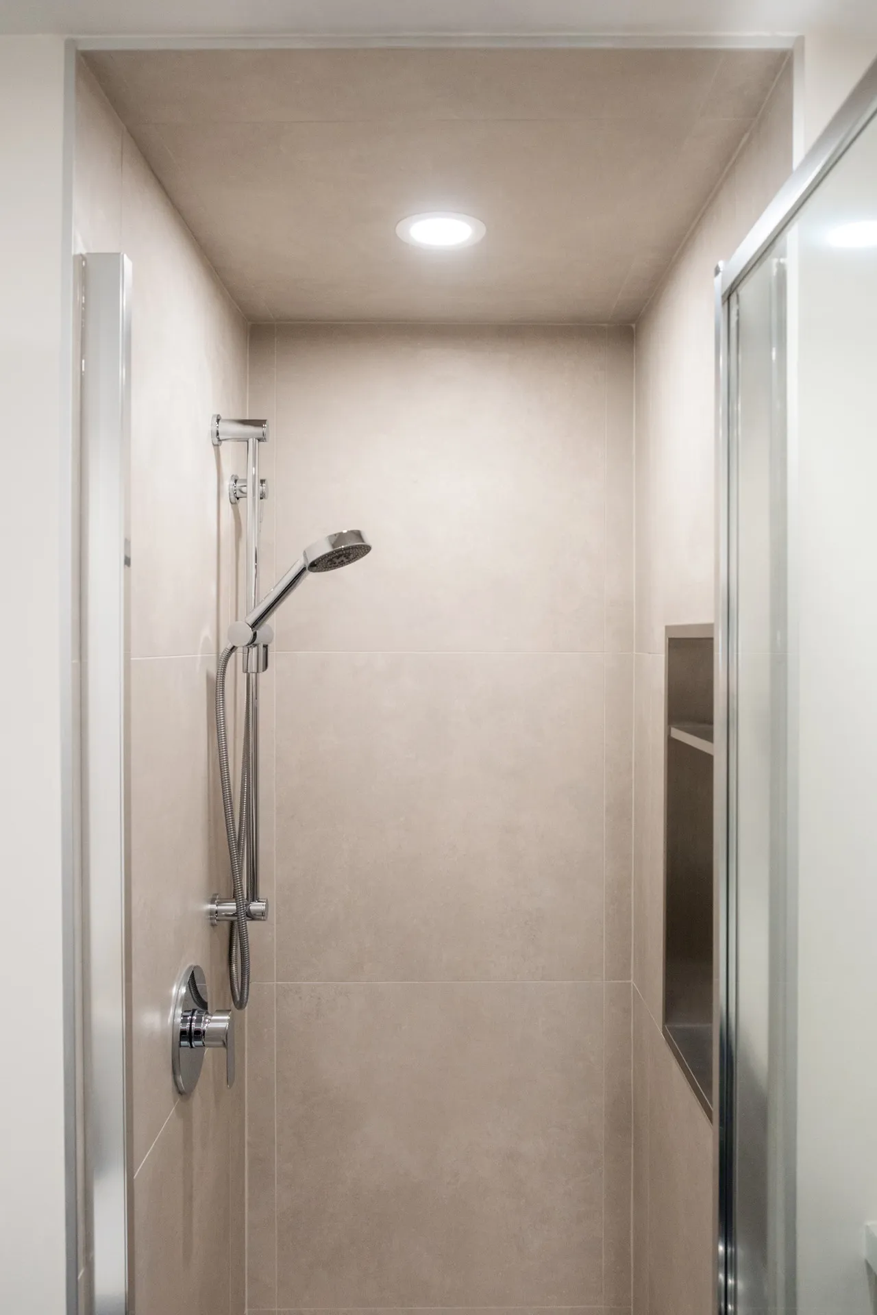 Douche moderne avec carrelage beige, mitigeur chromé et pommeau ajustable, éclairage encastré au plafond